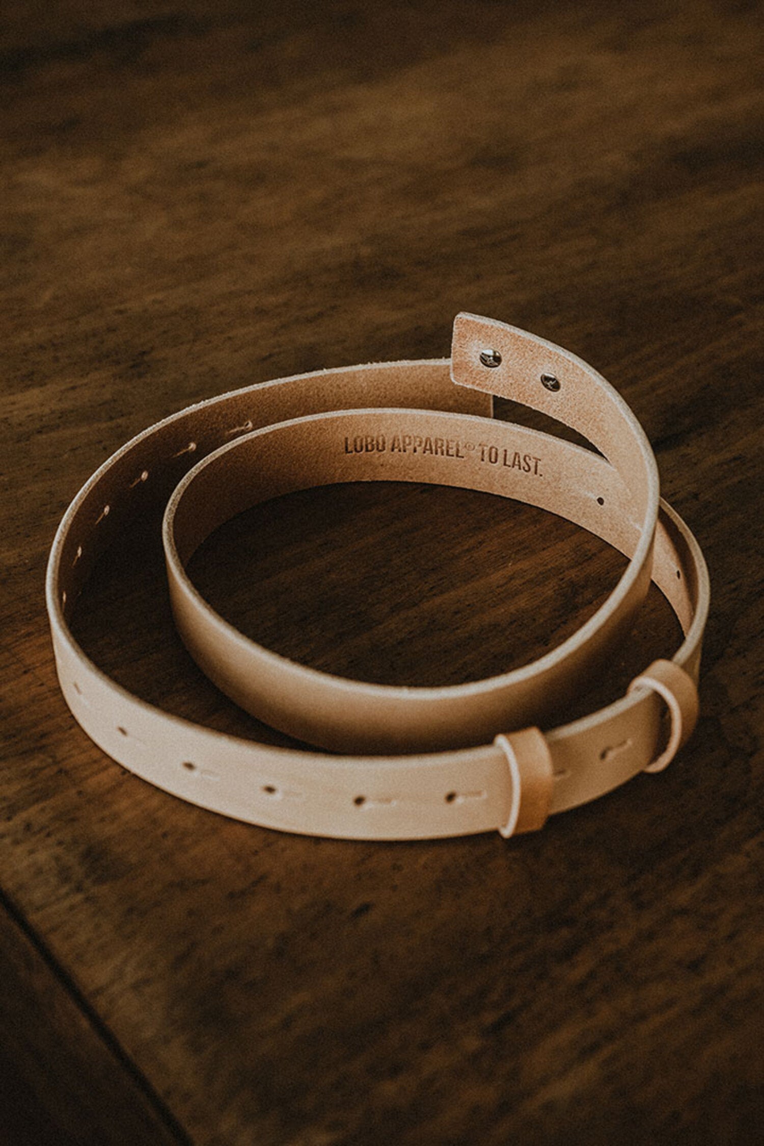 Tan genuine leather belt by Lobo Apparel, coiled, showing internal "LOBO APPAREL TO LAST" embossing on a wooden background.