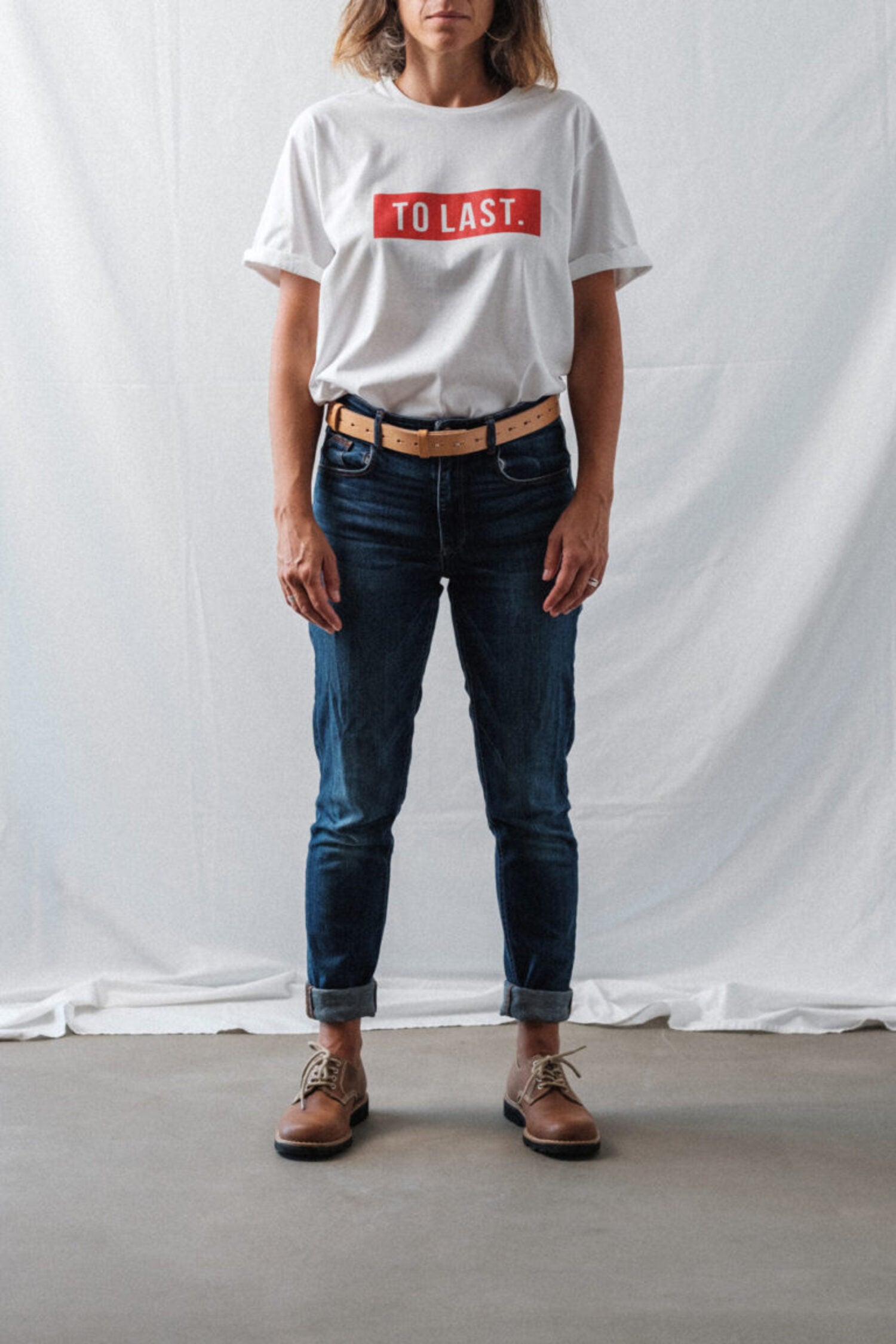 Woman wearing a "TO LAST" white t-shirt, blue jeans, and Lobo tan leather belt, embodying a minimalist and durable style.