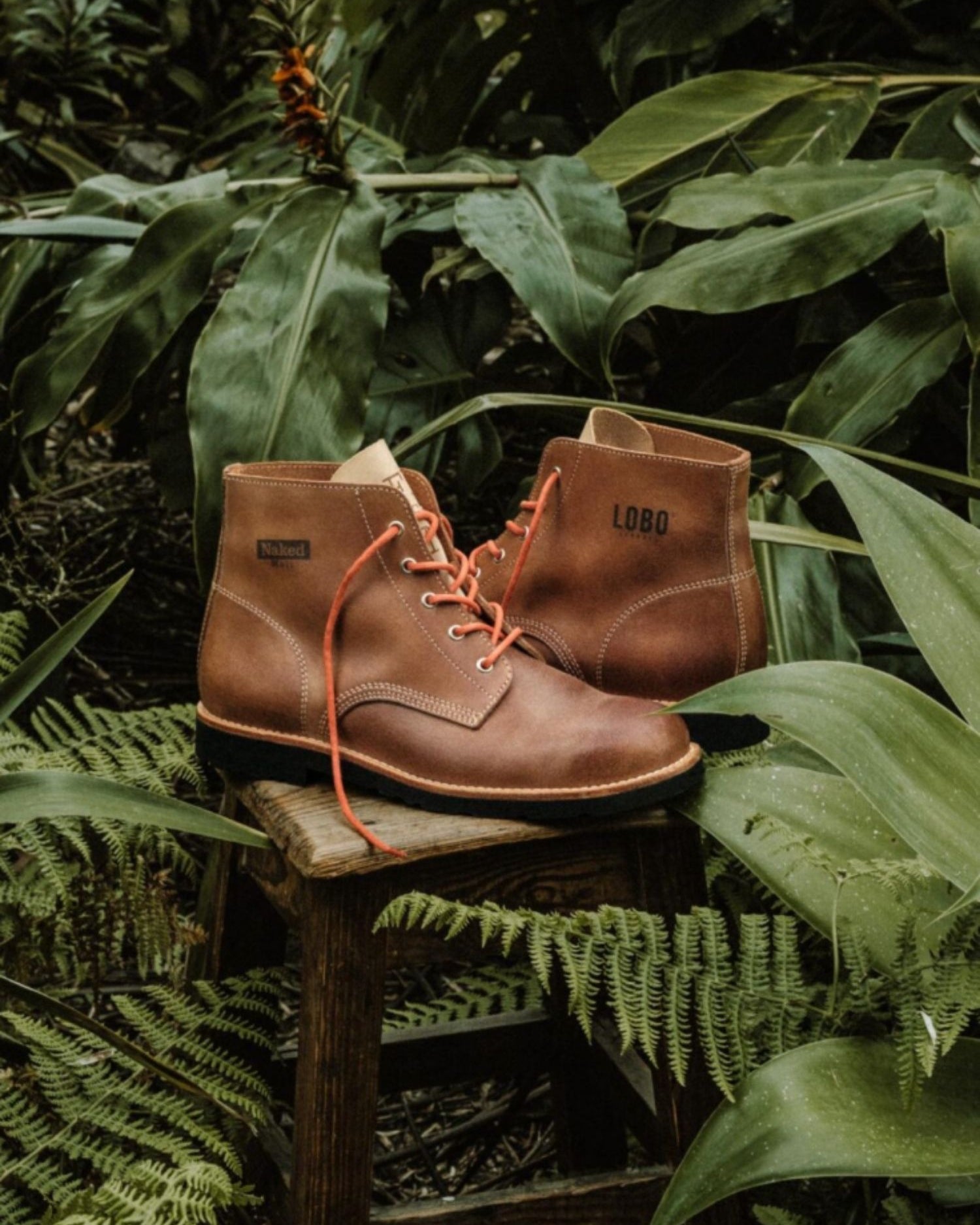 Handmade Lobo x Naked Malt Raw Boots in premium brown leather on a wooden stool surrounded by lush greenery.