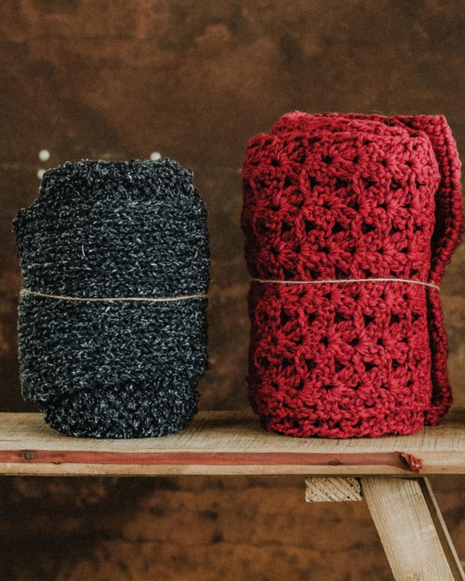 Two handmade Re-Born scarves — one in heather grey and another in burgundy crochet — crafted from repurposed materials, displayed on a wooden bench.