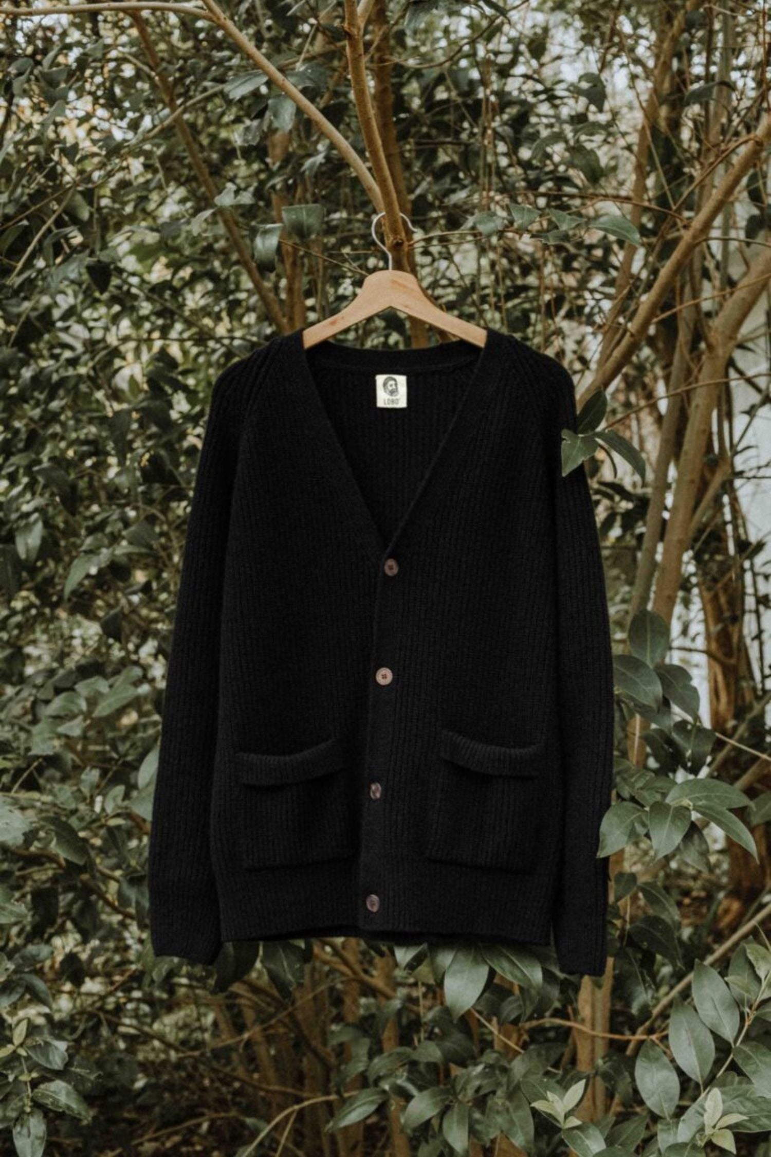 Full view of the black Lobo Black Wool Cardigan on a hanger, with front buttons and patch pockets in a natural setting