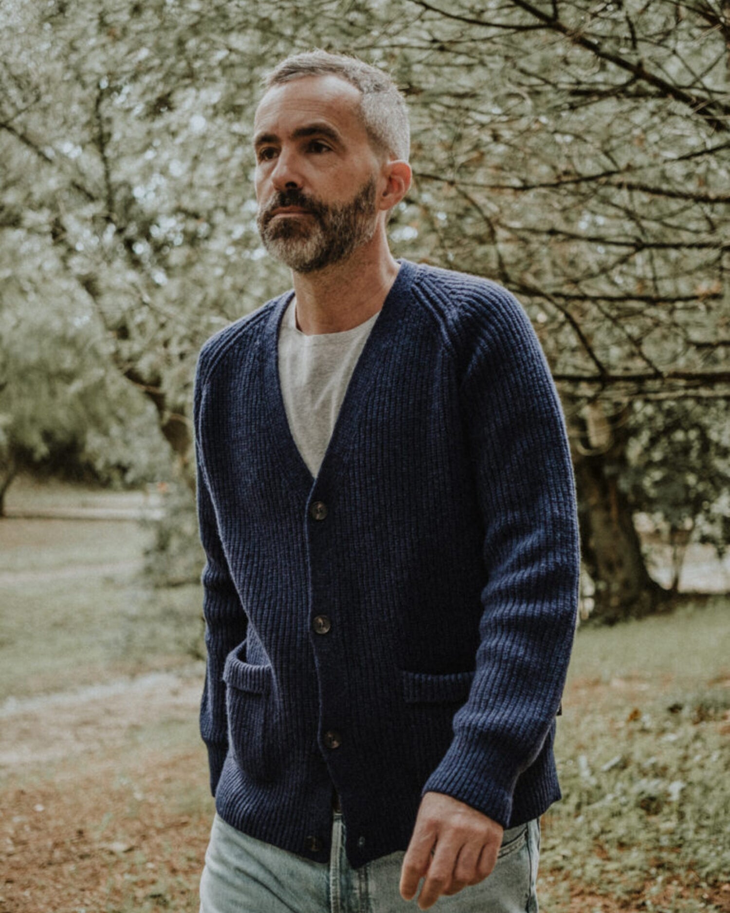 Man wearing the Lobo Apparel denim blue wool cardigan with a grey t-shirt, walking in an outdoor garden setting.