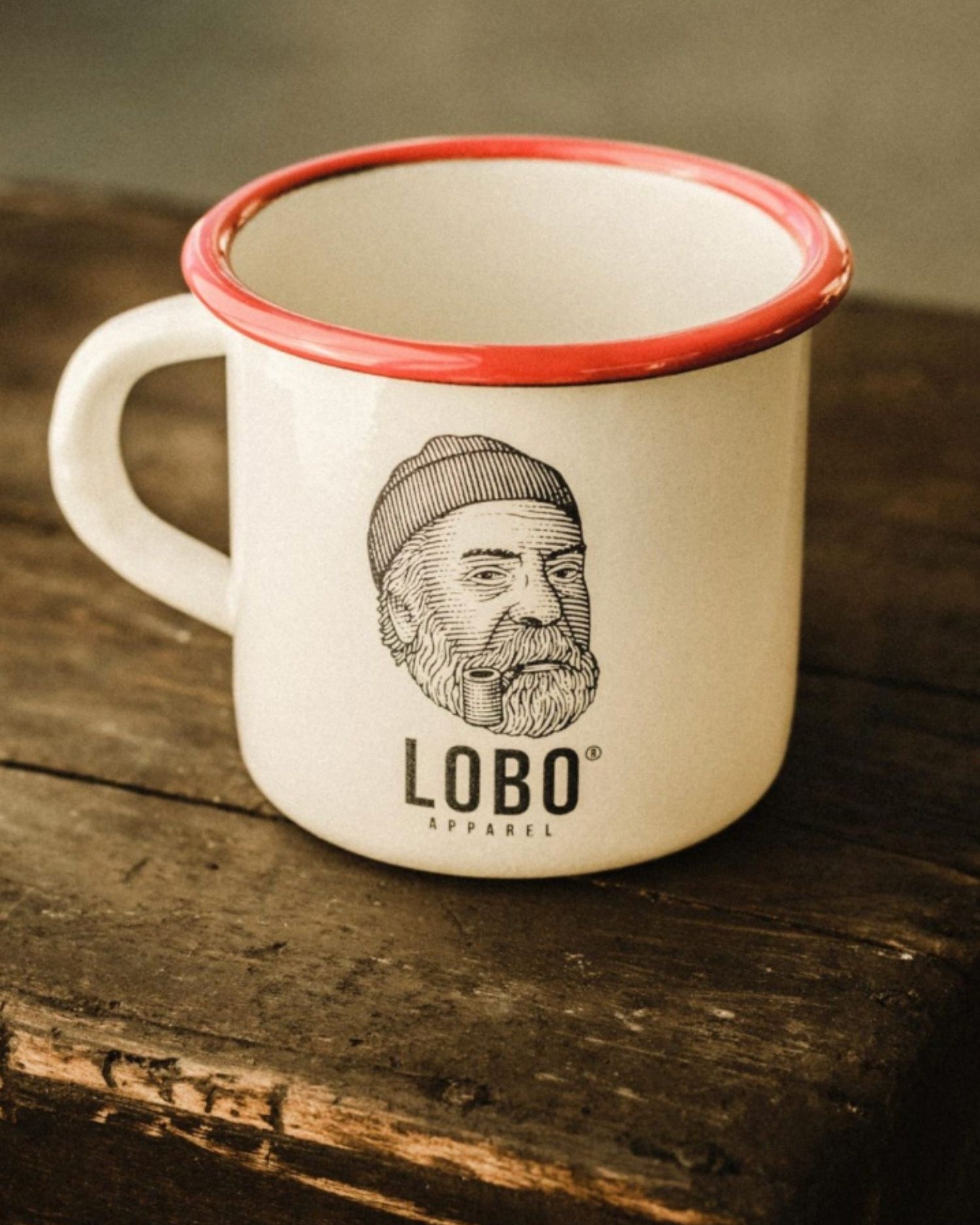 White enamel mug from LOBO Apparel with a red rim, featuring the illustration of a man wearing a beanie and smoking a pipe, set on an antique wooden box.