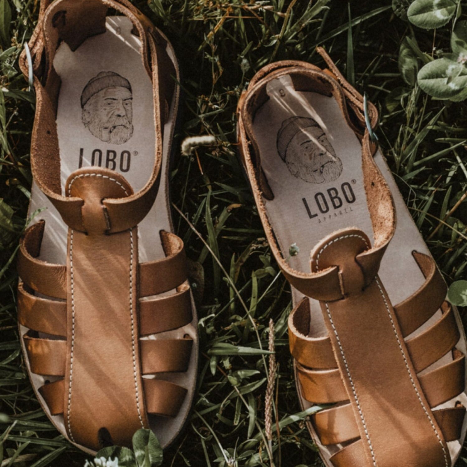Lobo brown leather sandals with rubber soles on green grass, featuring the leather insole with the brand's logo and illustration.