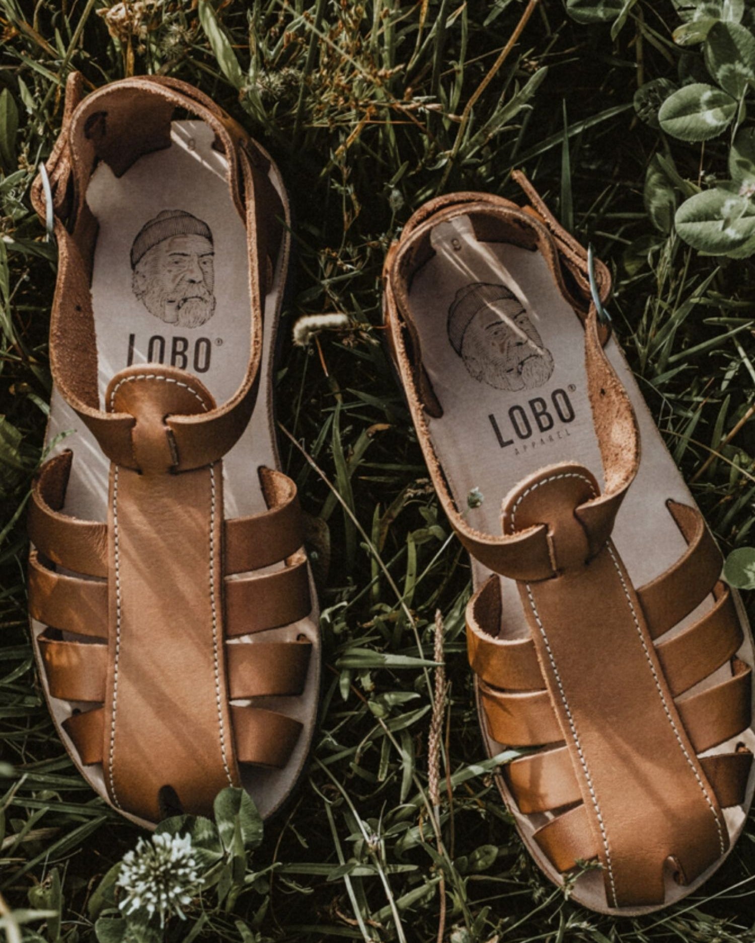 Lobo brown leather sandals with rubber soles on green grass, featuring the leather insole with the brand's logo and illustration.