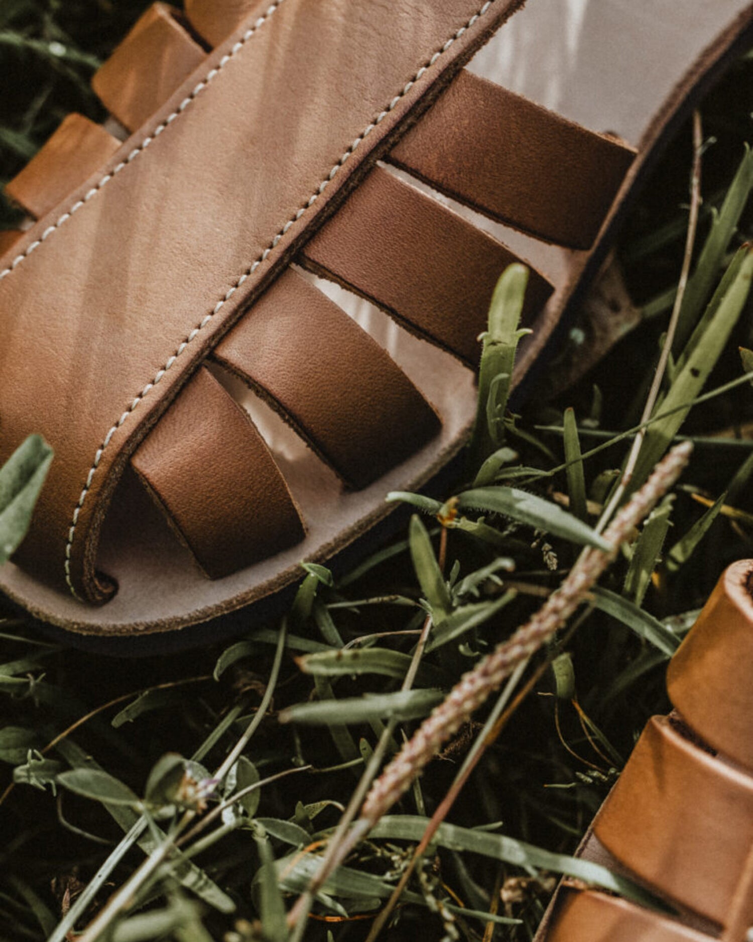 Close-up of Lobo brown leather sandals on grass, highlighting the detailed white stitching and the premium leather finish.