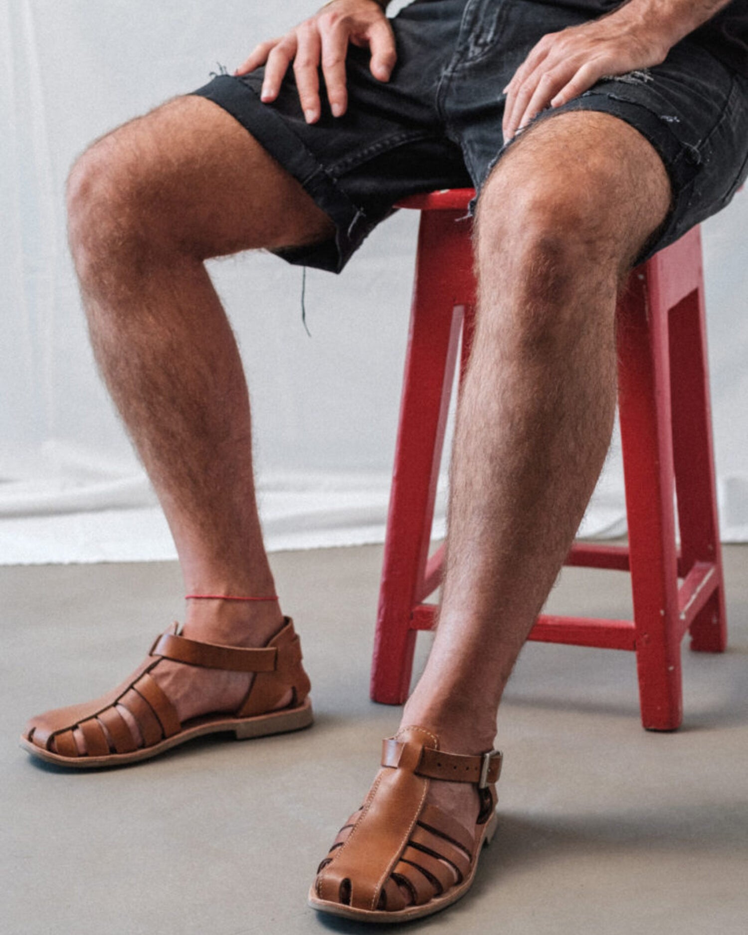 Man sitting on a red stool wearing brown Lobo Leather Sandals, demonstrating the comfortable fit and minimalist leather strap design.