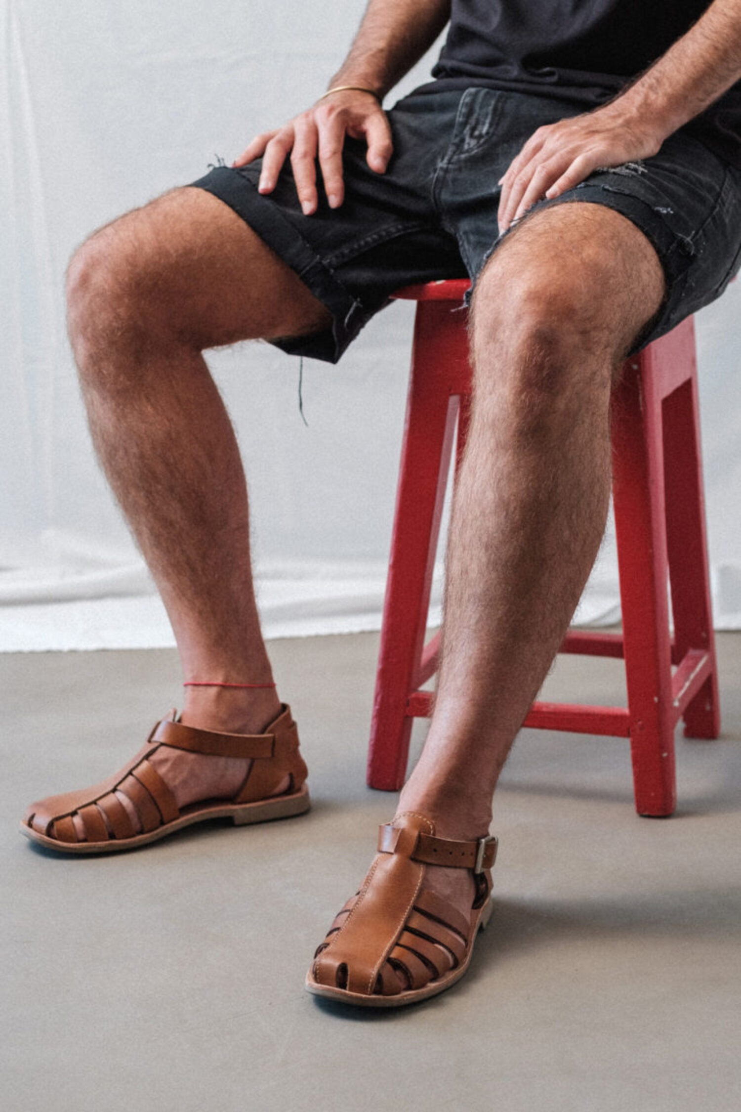 Man sitting on a red stool wearing brown Lobo Leather Sandals, demonstrating the comfortable fit and minimalist leather strap design.