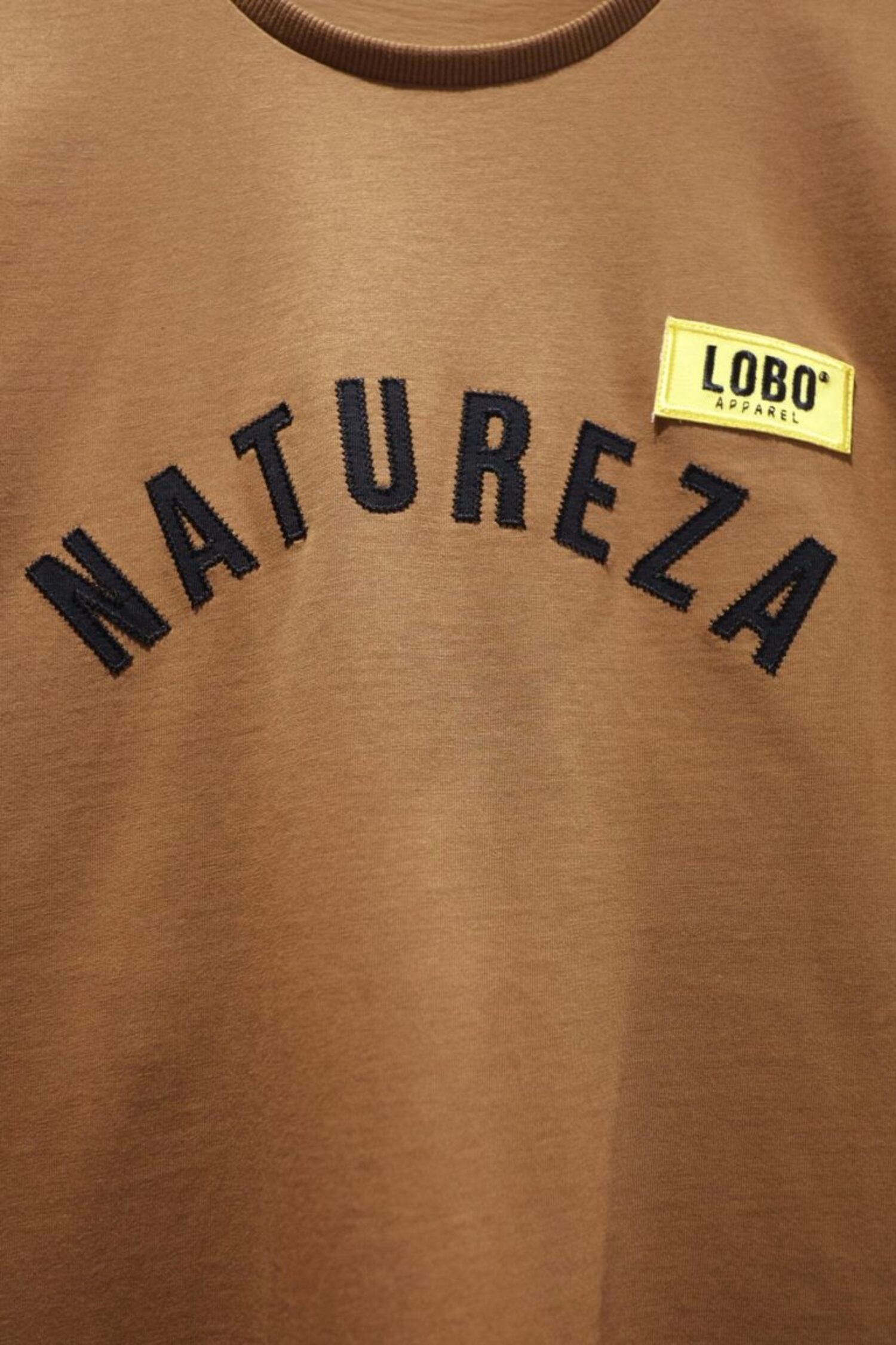 Detail of the black arched embroidery with the word "NATUREZA" and a yellow sewn patch on a premium brown cotton t-shirt.