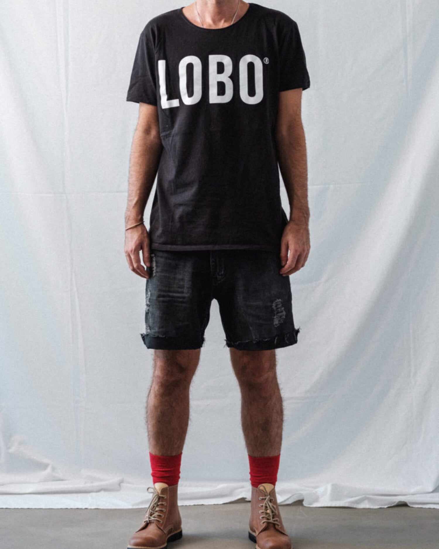 Full-body shot of a male model wearing a black Lobo Apparel logo t-shirt, distressed black denim shorts, and brown leather boots with red socks against a white backdrop.