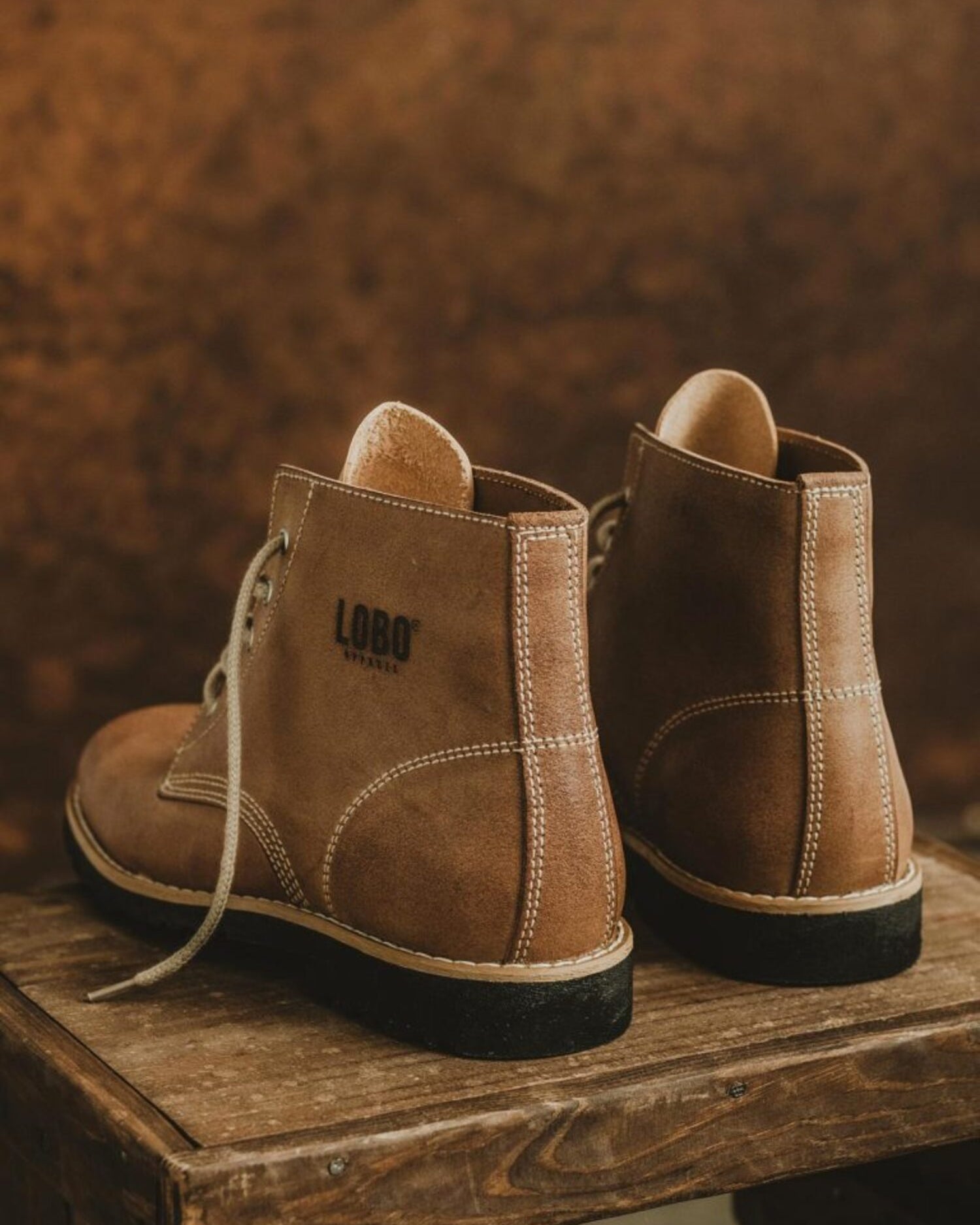 Rear view of Lobo Raw brown leather boots on a rustic wooden stool, highlighting the branded logo stamp.
