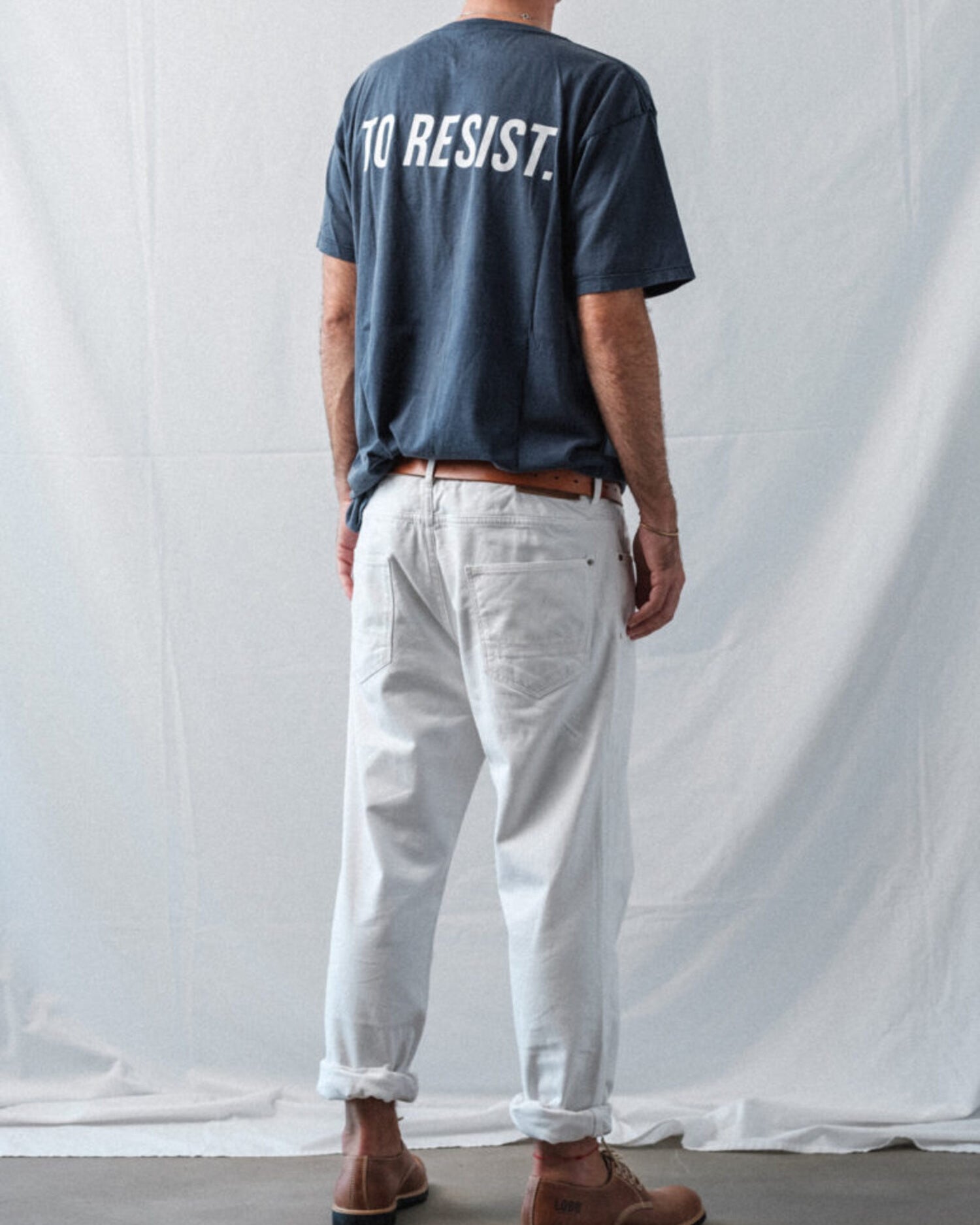 Back view of a man wearing Lobo Raw shoes, white trousers, and a blue t-shirt with "To Resist" text.