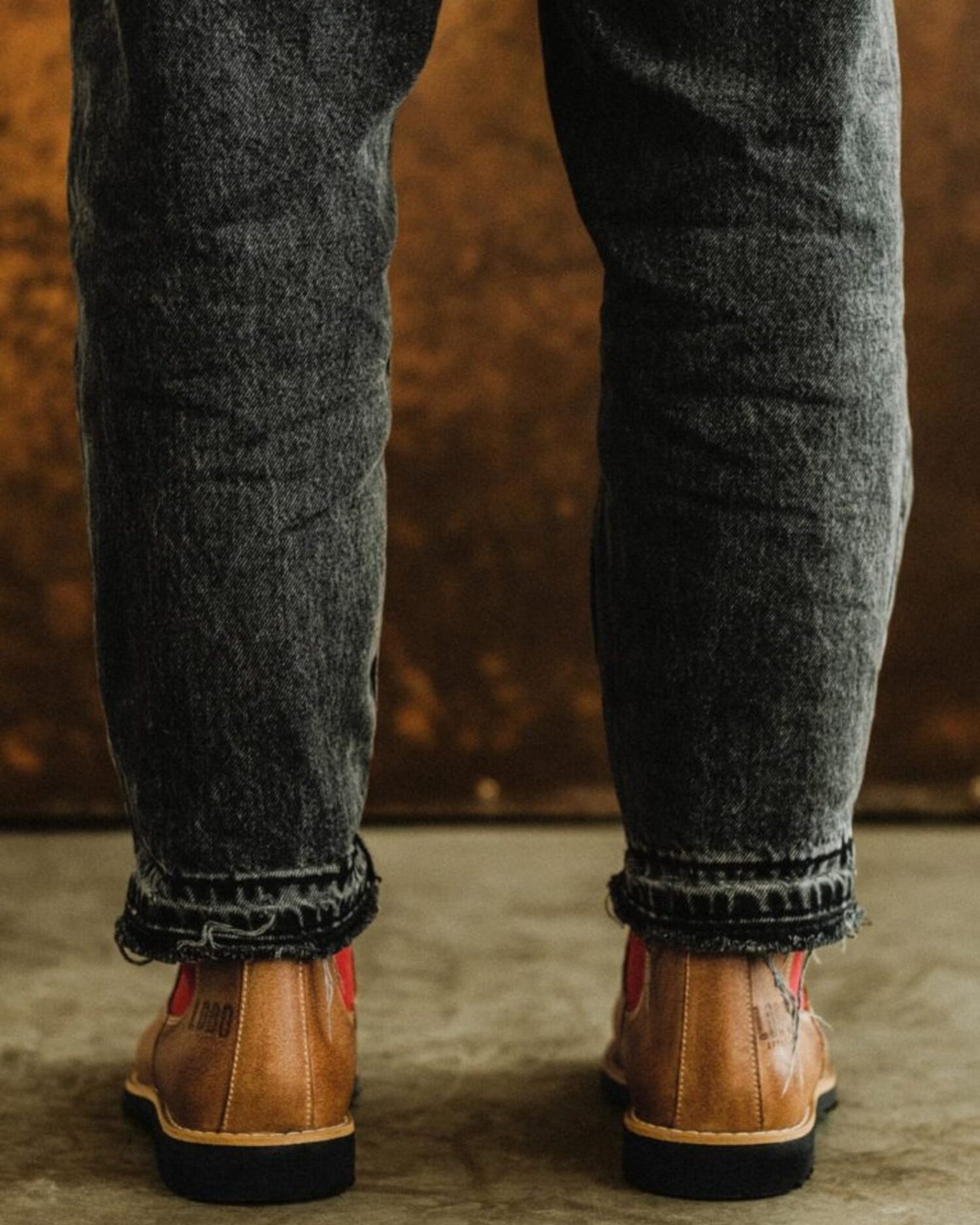 Rear view of a person wearing Lobo tan leather boots with dark wash jeans, showing red side panels.