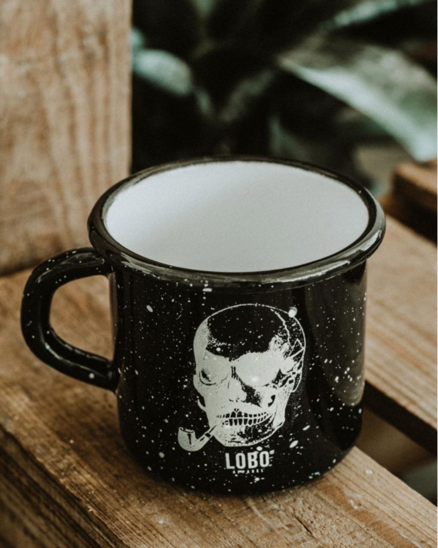 Lobo Skull black enamel mug featuring a skull smoking a pipe illustration, resting on rustic wood.