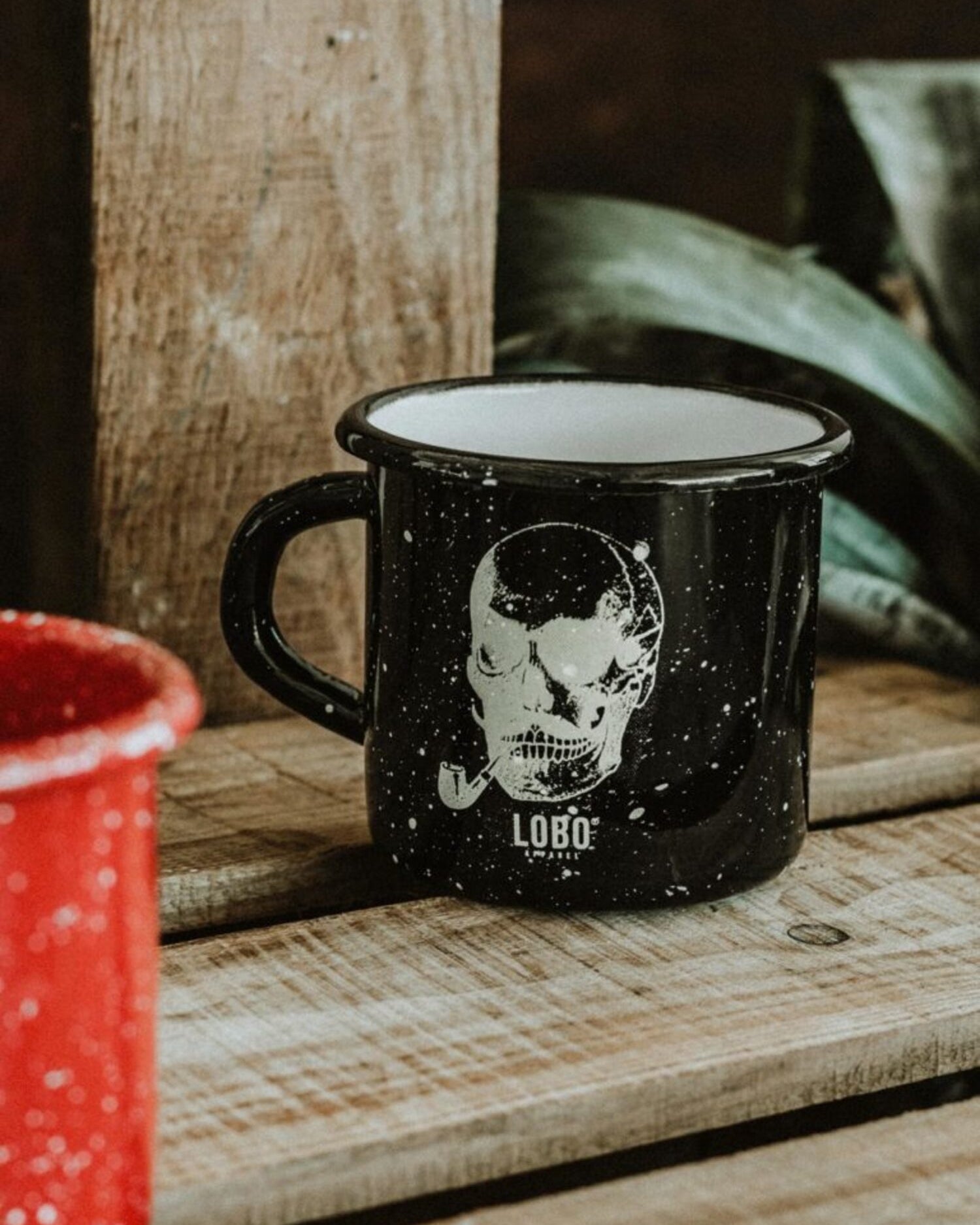 Black Lobo Skull Enamel mug with skull illustration on a wooden surface, featuring a partial red mug and plant in the background.