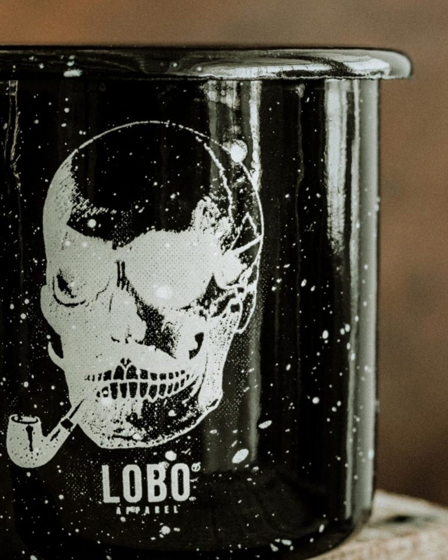 Close-up of the black Lobo Skull Enamel mug, highlighting the speckled enamel texture and the detailed illustration of a skull with a pipe.