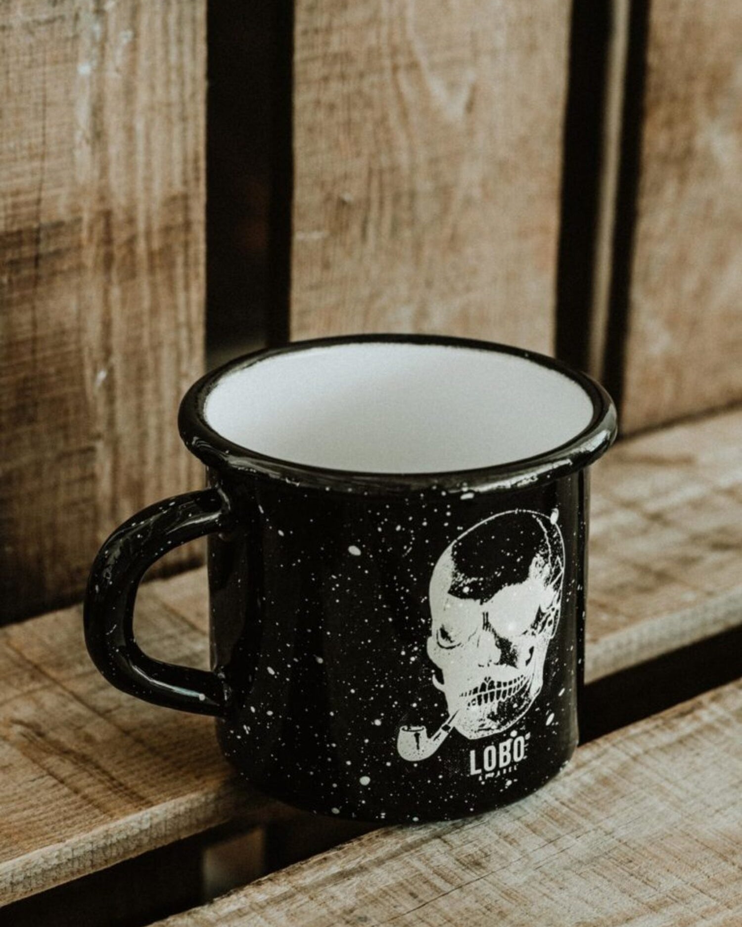 Black Lobo Skull enamel metal mug with white interior, featuring a skull illustration and logo on rustic wooden planks.