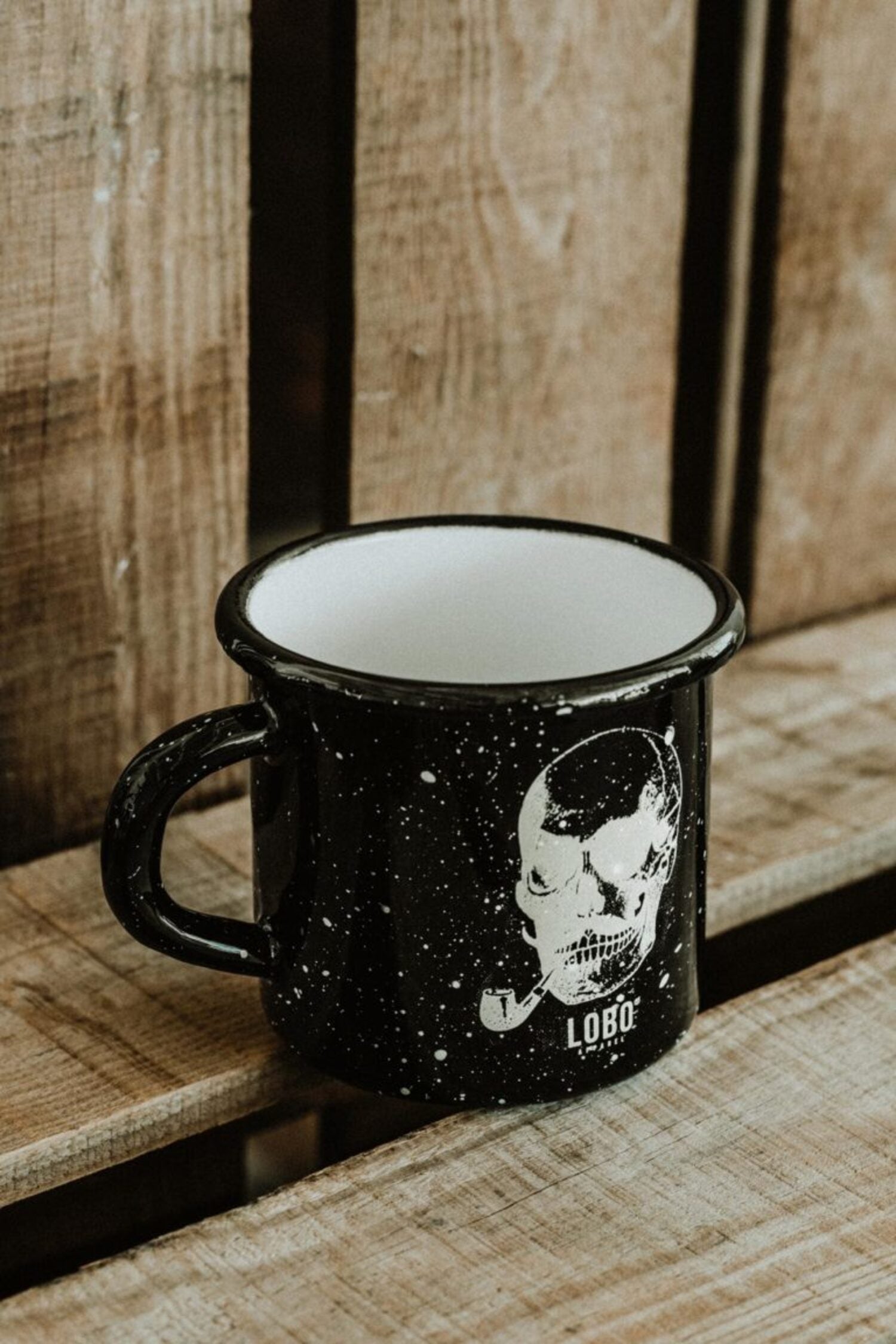 Black Lobo Skull enamel metal mug with white interior, featuring a skull illustration and logo on rustic wooden planks.