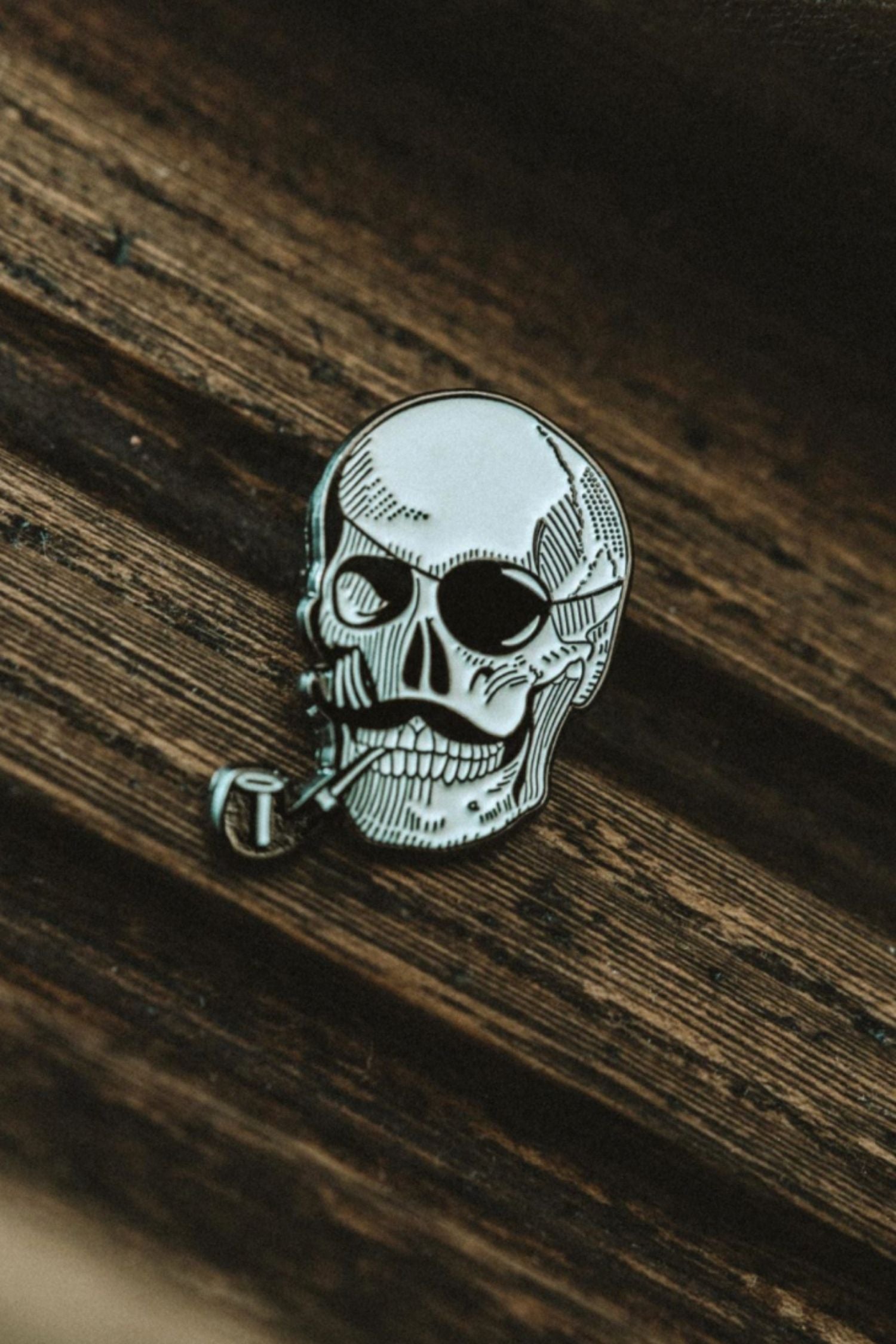 Lobo Skull metal pin featuring a detailed skull design with a mustache and pipe on a dark wood surface.