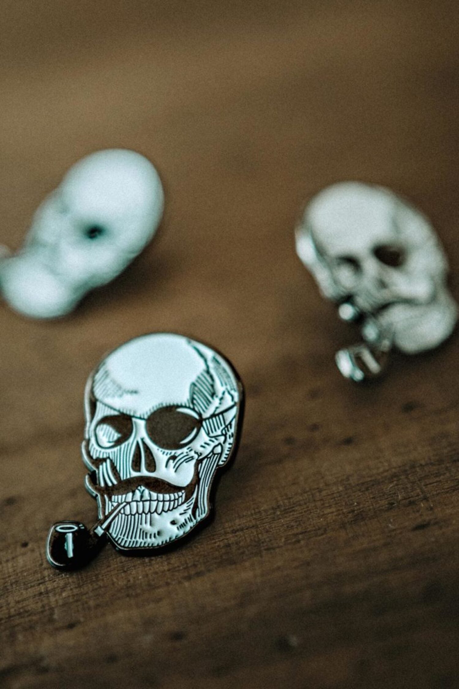 Artistic close-up of the Lobo Skull metal pin featuring a skull with a mustache and pipe design, focused on a rustic wooden surface.