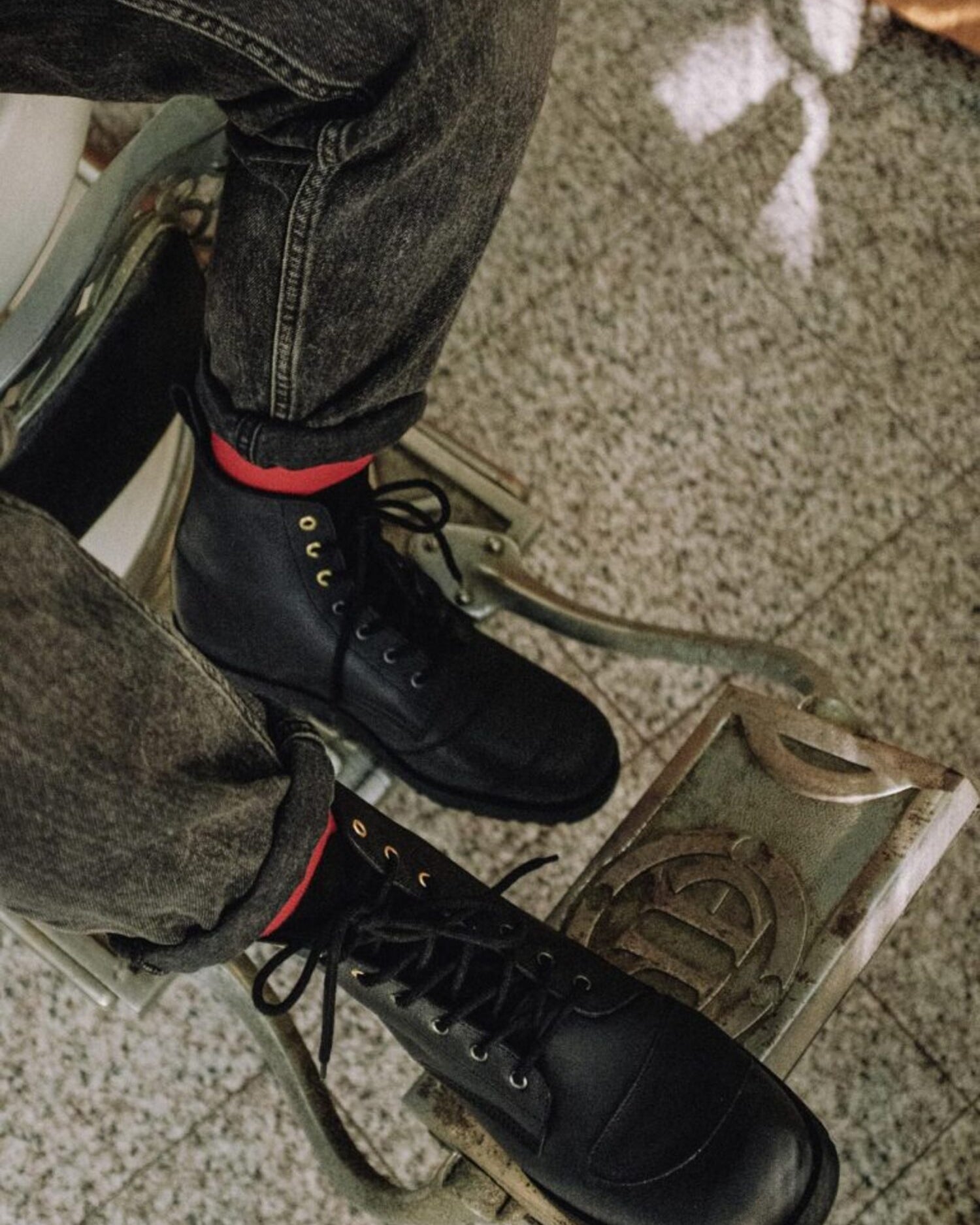 High-angle view of the black leather Lobo Tigre Boots 3rd Edition, worn with red socks and grey denim jeans, highlighting the golden eyelets and the motorcycle shifter protector.
