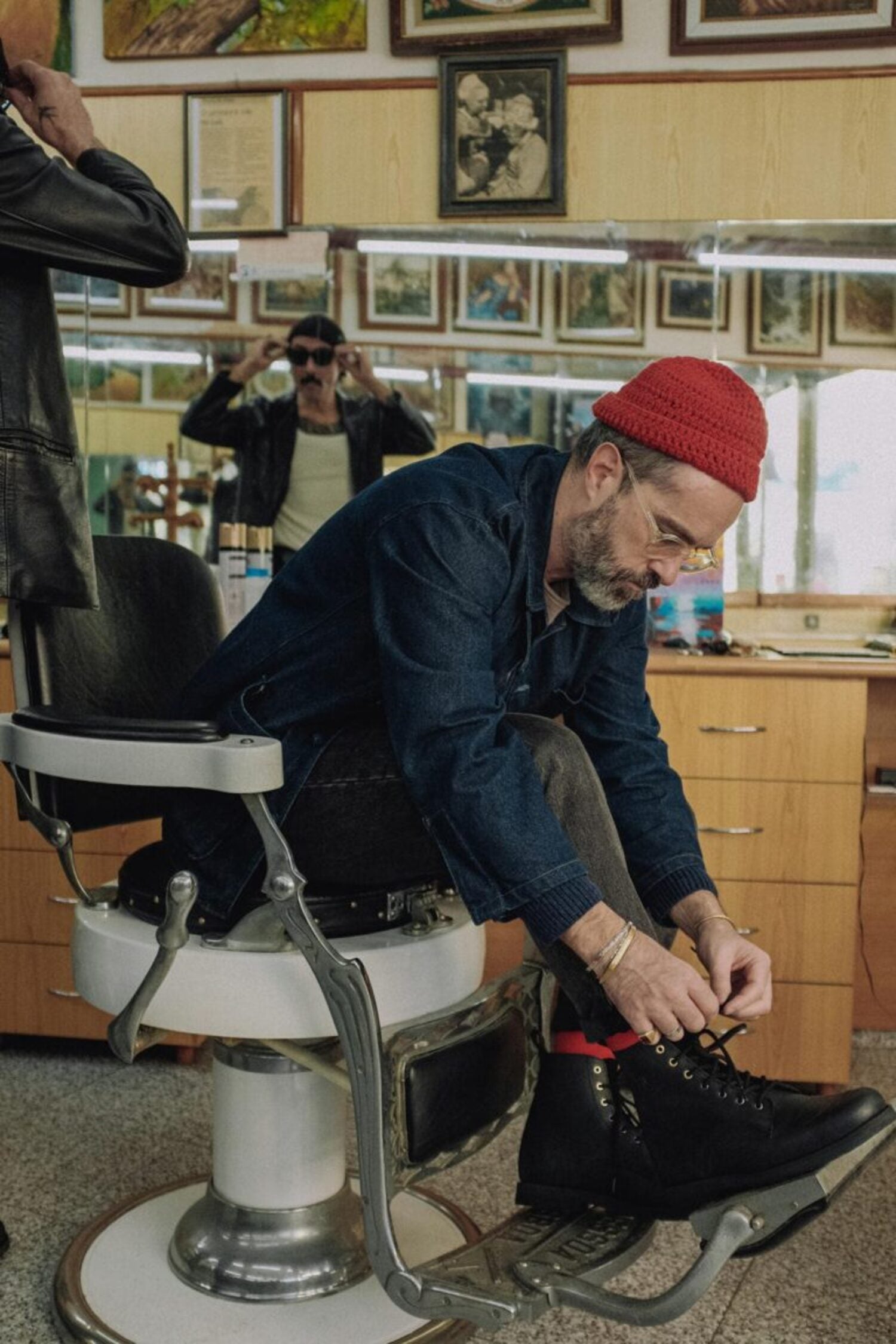 Man sitting in a classic barbershop chair putting on the Lobo Tigre Boots 3rd Edition, demonstrating the fit and rugged style of the footwear.