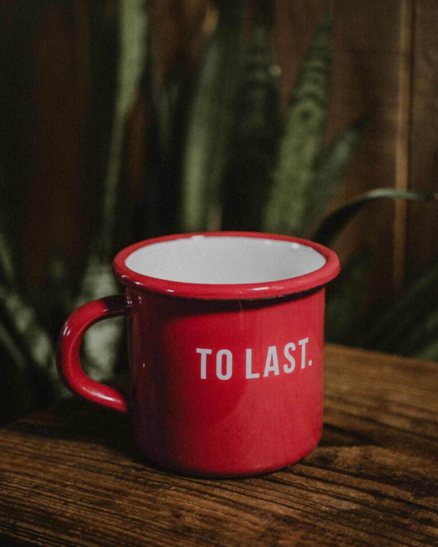 "To Last" red enamel mug on a rustic wooden table with a plant in the background.
