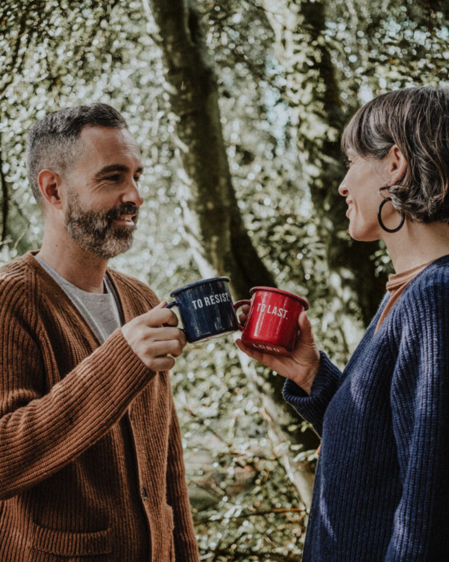 Couple toasting with Lobo "To Resist" blue and "To Last" red enamel mugs in a forest setting.