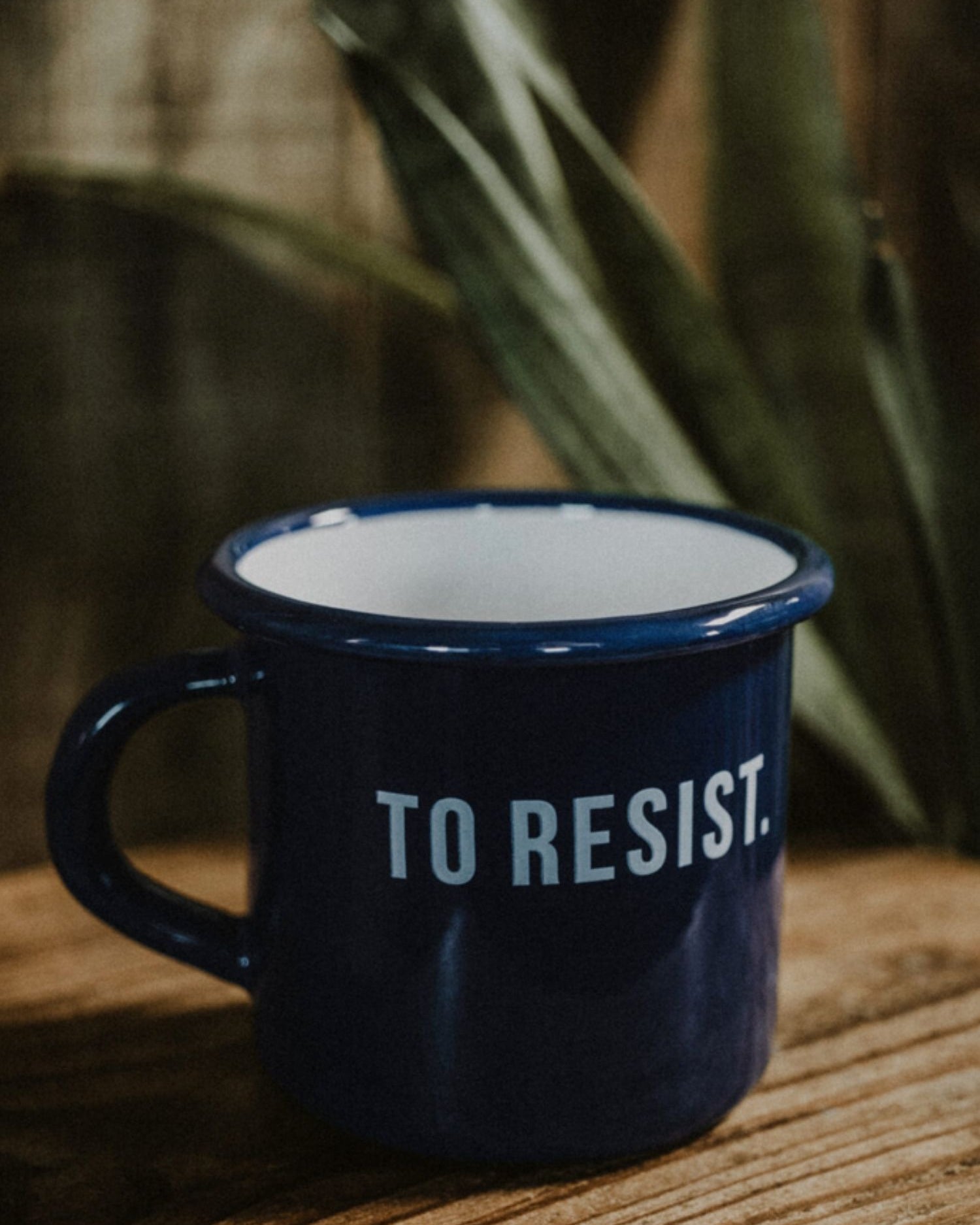 Lobo 'To Resist' navy blue enamel mug, 0.40 L capacity, displayed on a rustic wooden table with plants in the background.