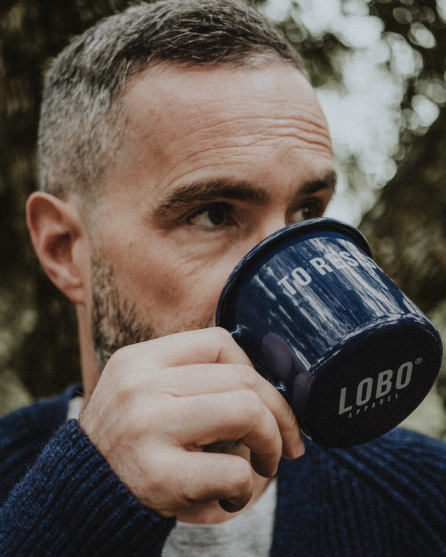 Close-up of a bearded man drinking from the blue 'To Resist' enamel mug, highlighting the Lobo Apparel logo on the base, in a nature setting.
