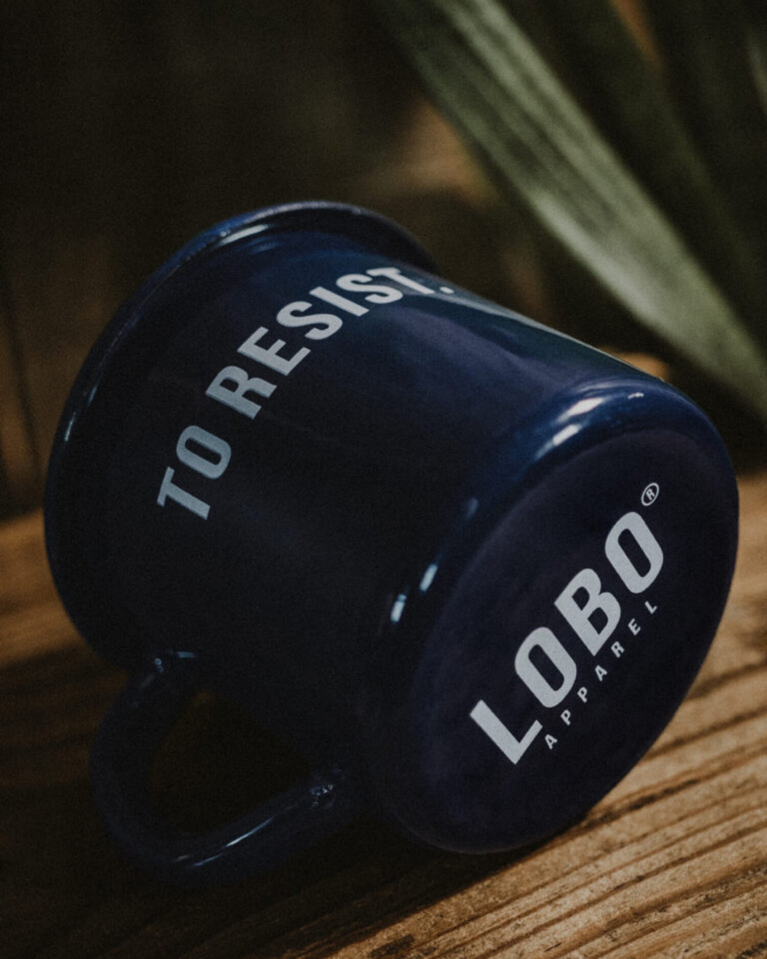 Blue 'To Resist' enamel mug lying on wood, highlighting the Lobo Apparel logo on the base and its glossy finish.