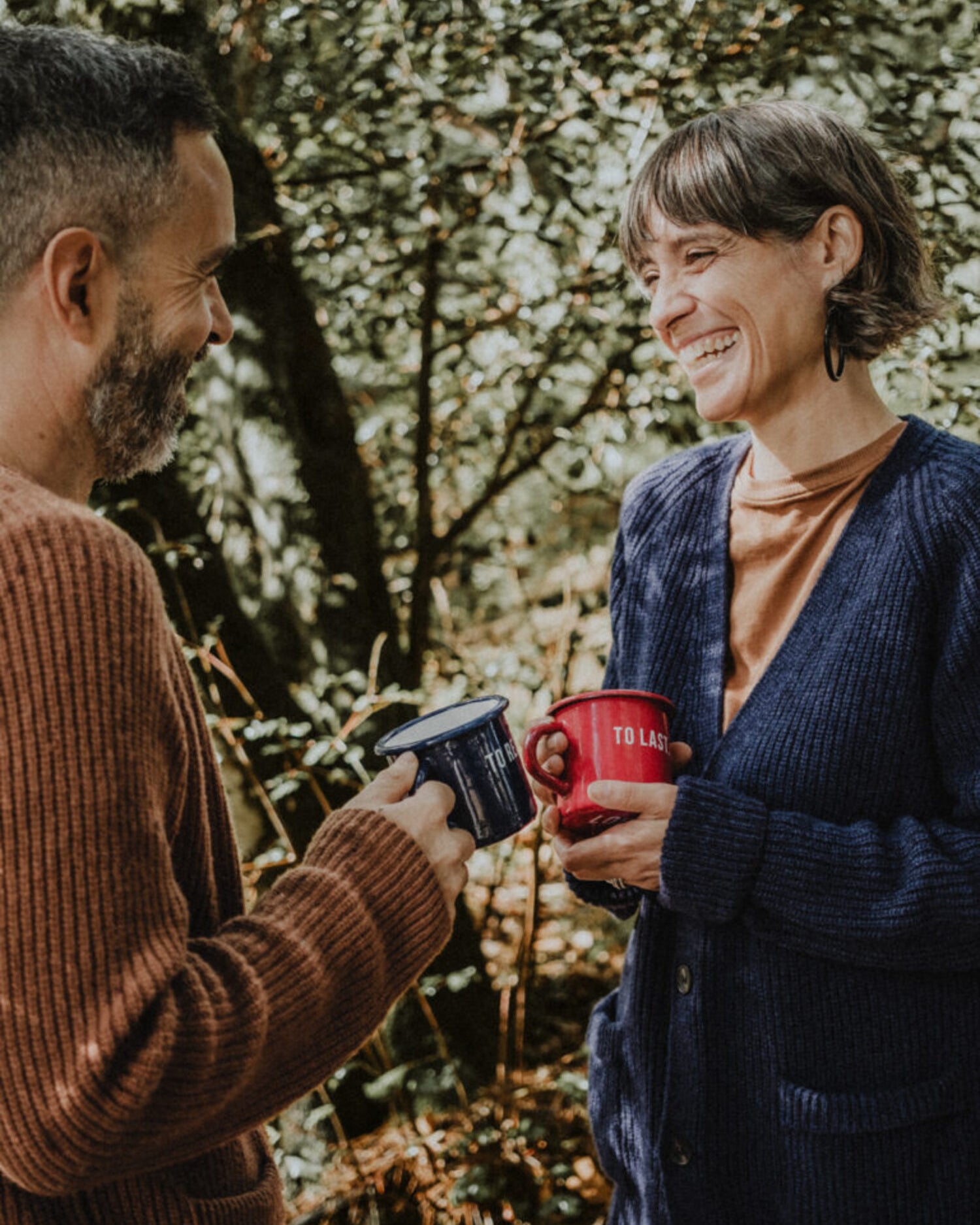 Smiling man and woman in a forest setting, holding Lobo 'To Resist' and 'To Last' enamel mugs, wearing cozy wool knitwear.