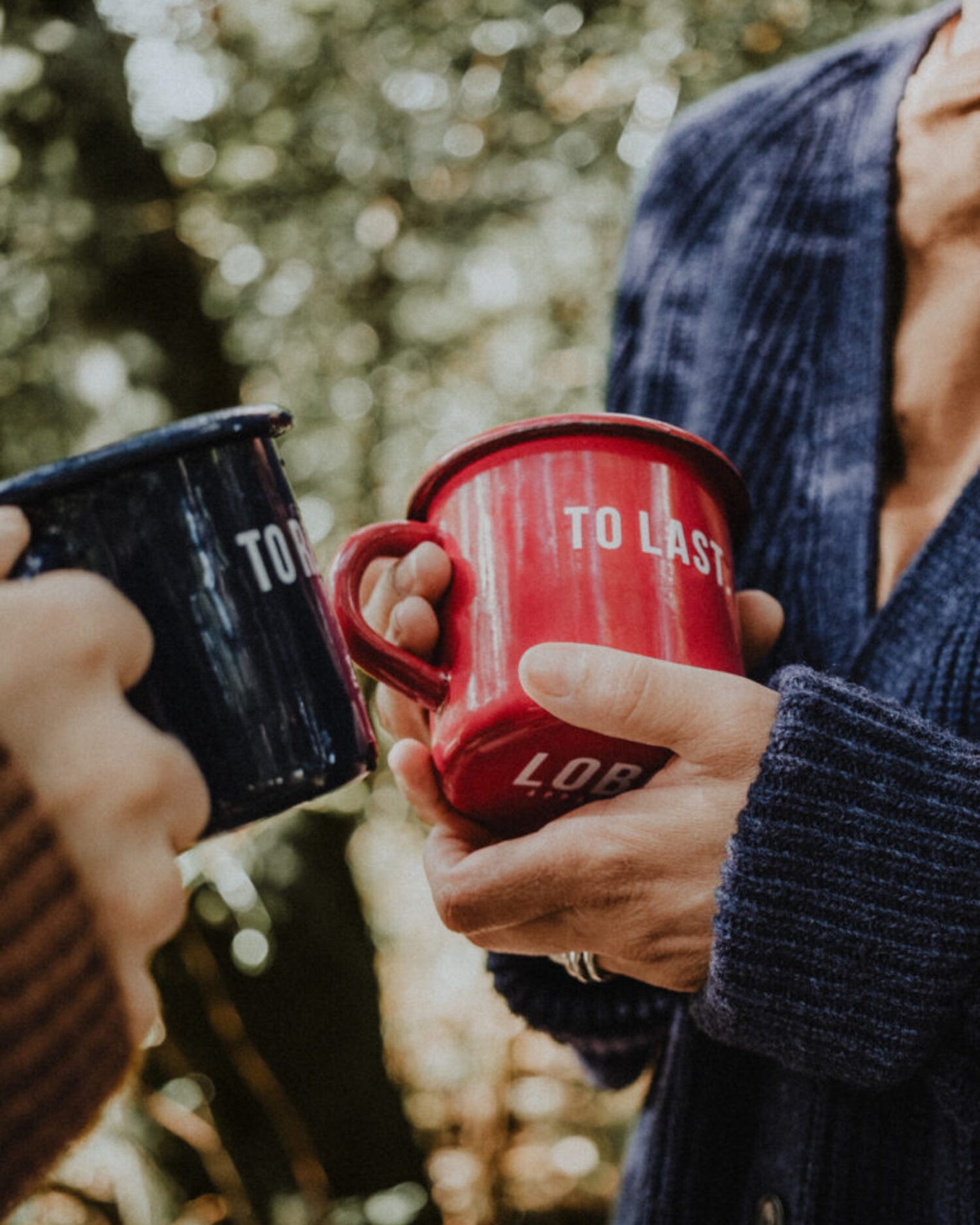 Two people holding Lobo enamel mugs — 'To Resist' in blue and 'To Last' in red — in an outdoor forest setting.