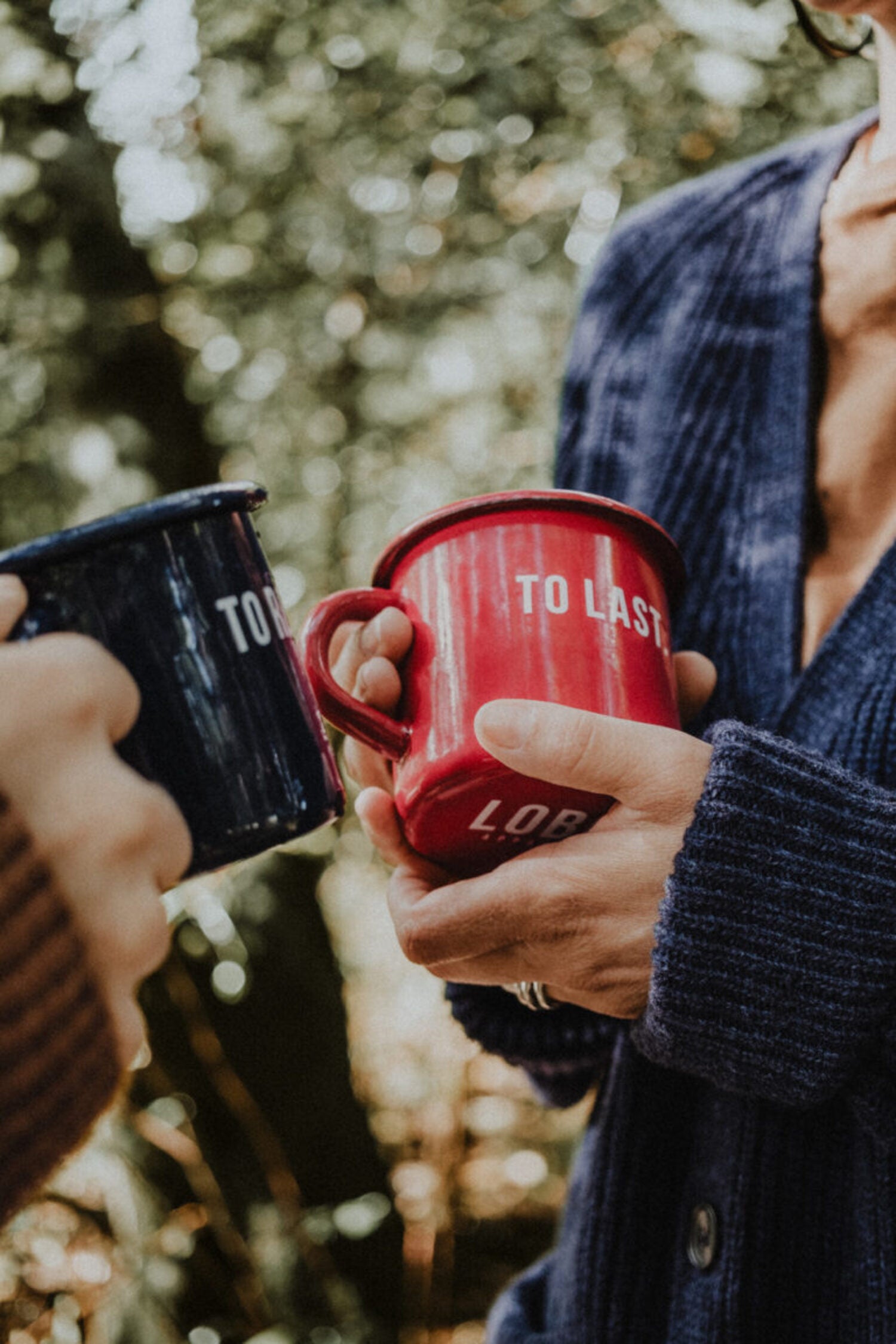 Two people holding Lobo enamel mugs — 'To Resist' in blue and 'To Last' in red — in an outdoor forest setting.