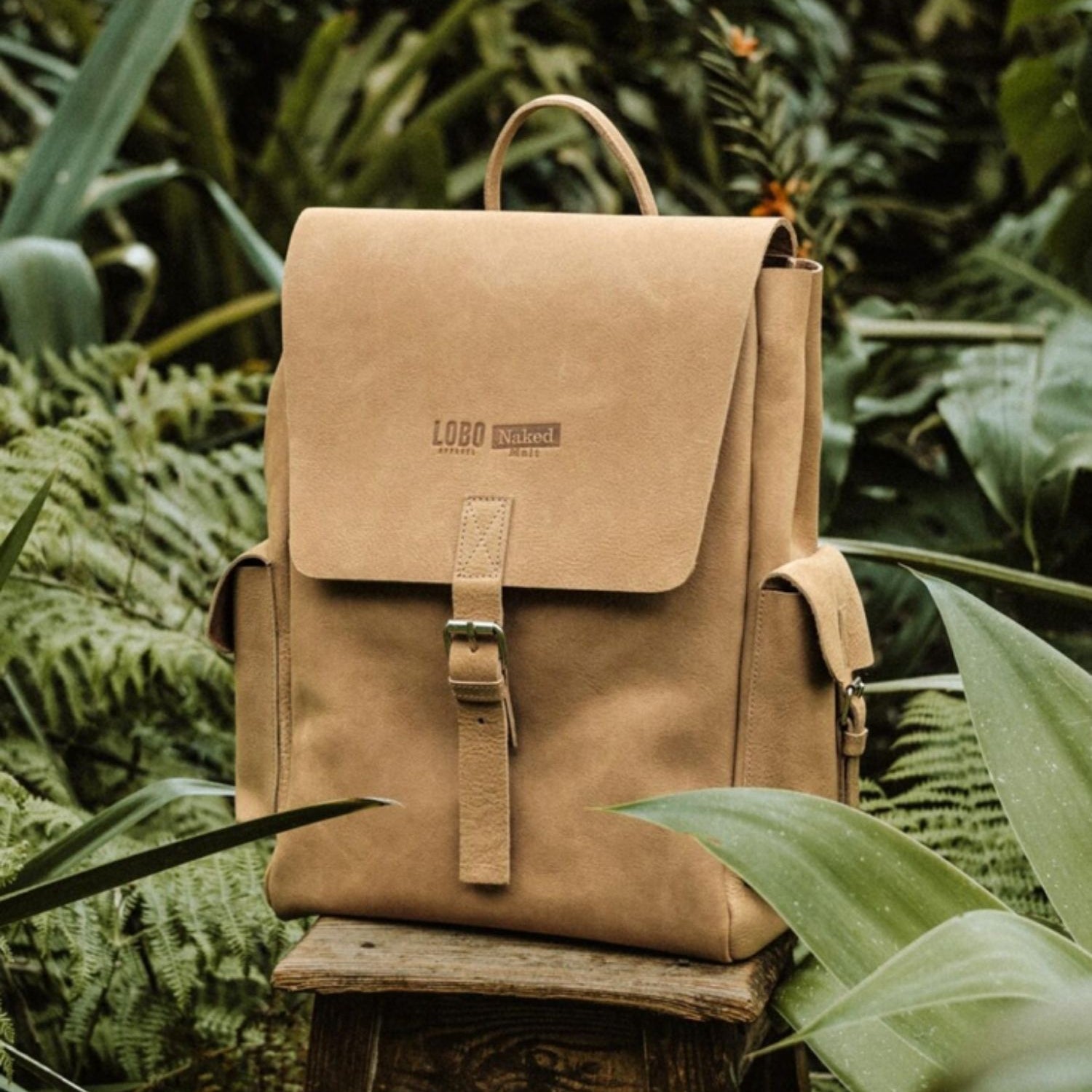 Lobo Apparel x Naked Malt brown leather backpack, minimalist design with center buckle, placed on a wooden stool in a natural outdoor setting.