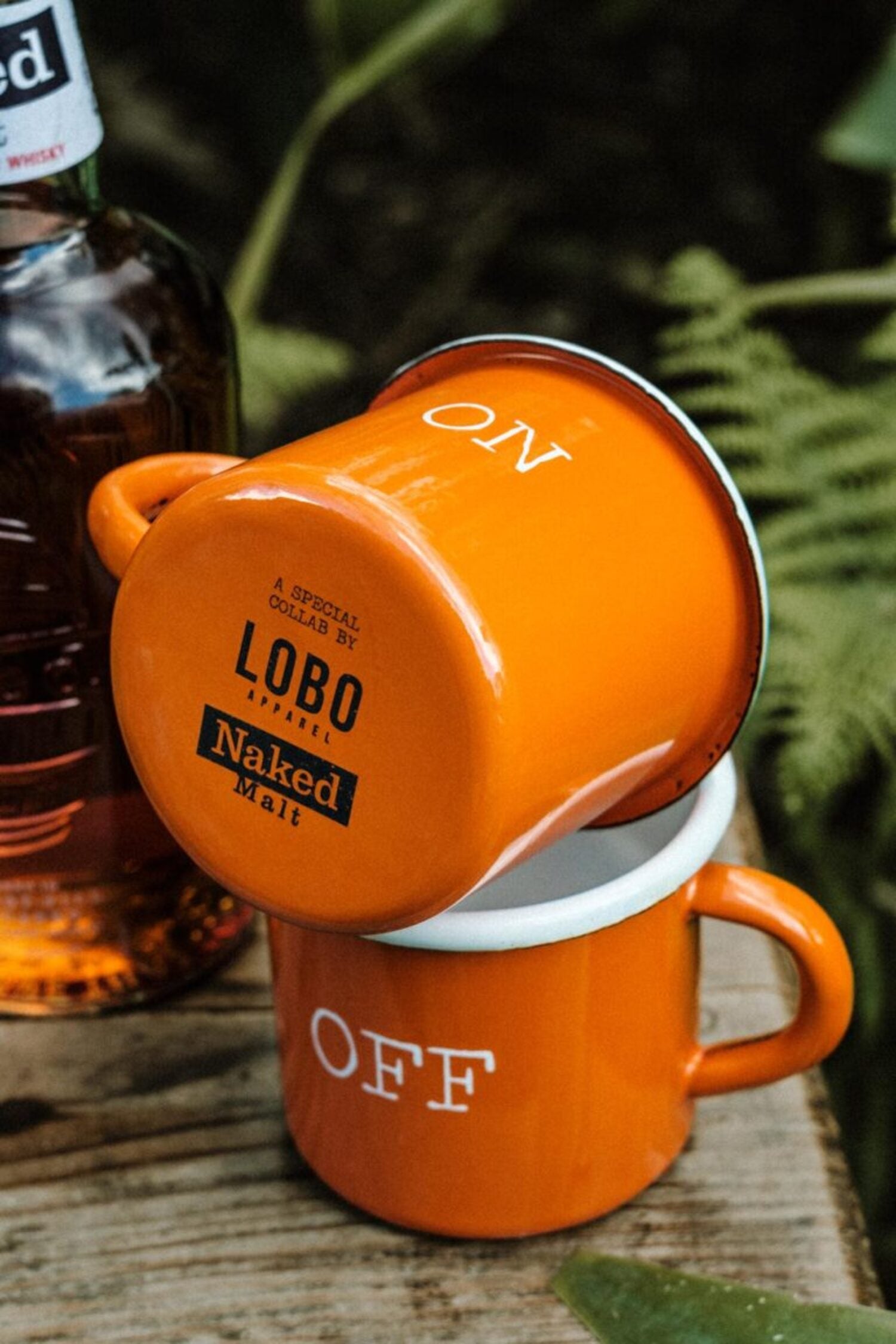 Close-up of Lobo Apparel x Naked Malt mug base featuring collaboration logos, stacked on an "OFF" mug in a rustic setting.