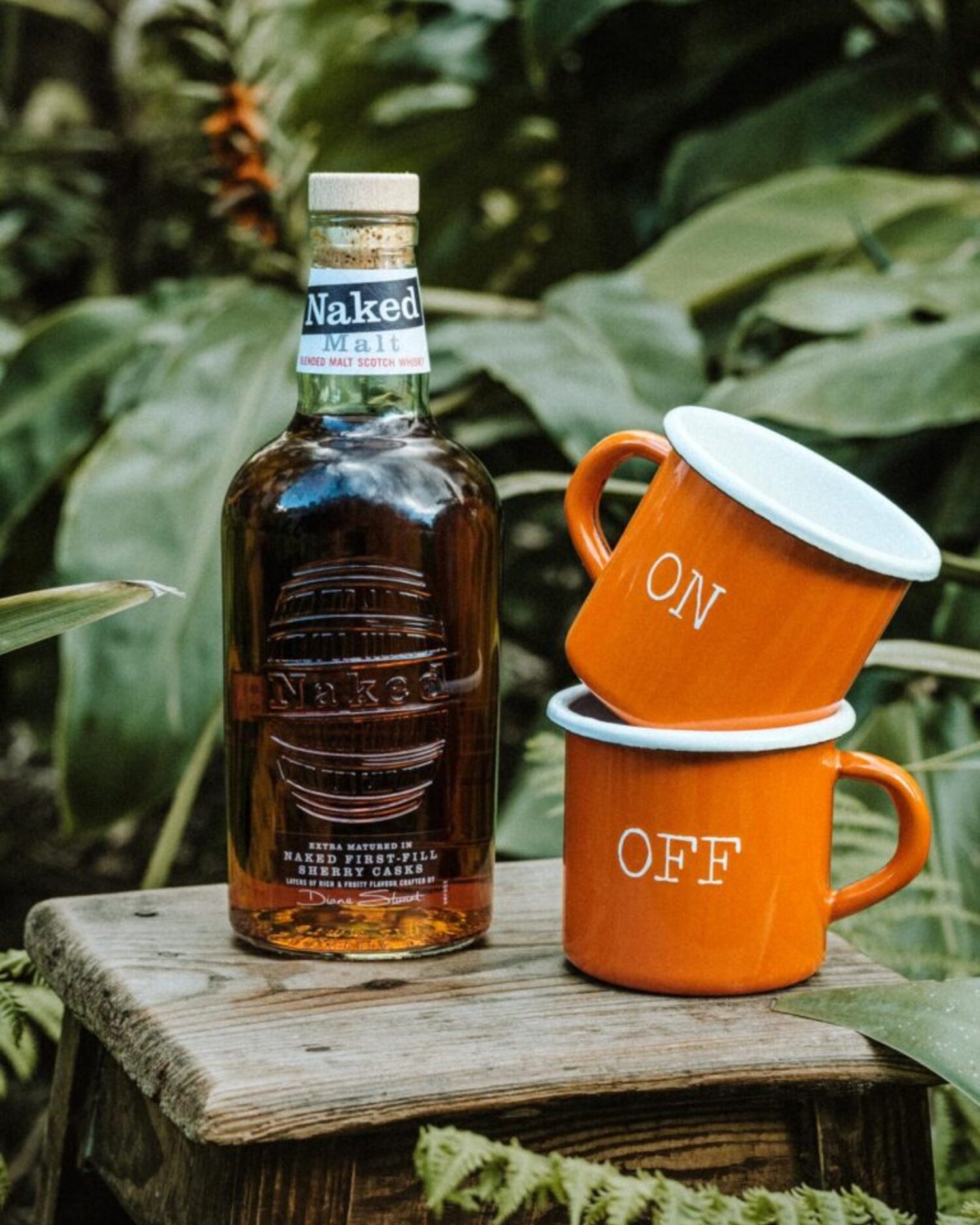 Lobo Apparel ON OFF mugs next to a Naked Malt whisky bottle in a tropical forest setting.