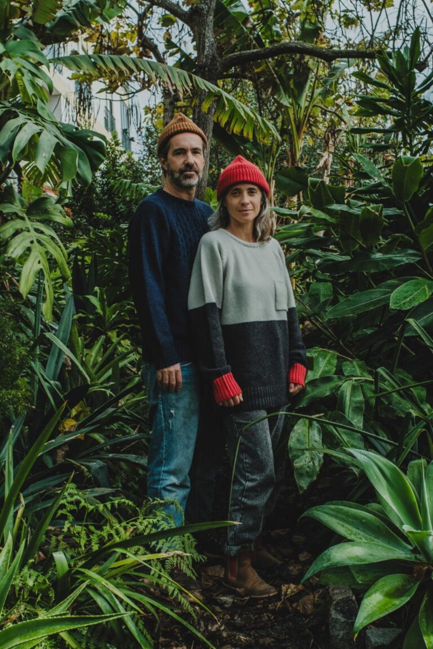 A couple in a lush tropical garden; the man wears the Manuel Brown Beanie wool hat and a blue knit sweater, while the woman wears a red beanie and two-tone sweater.