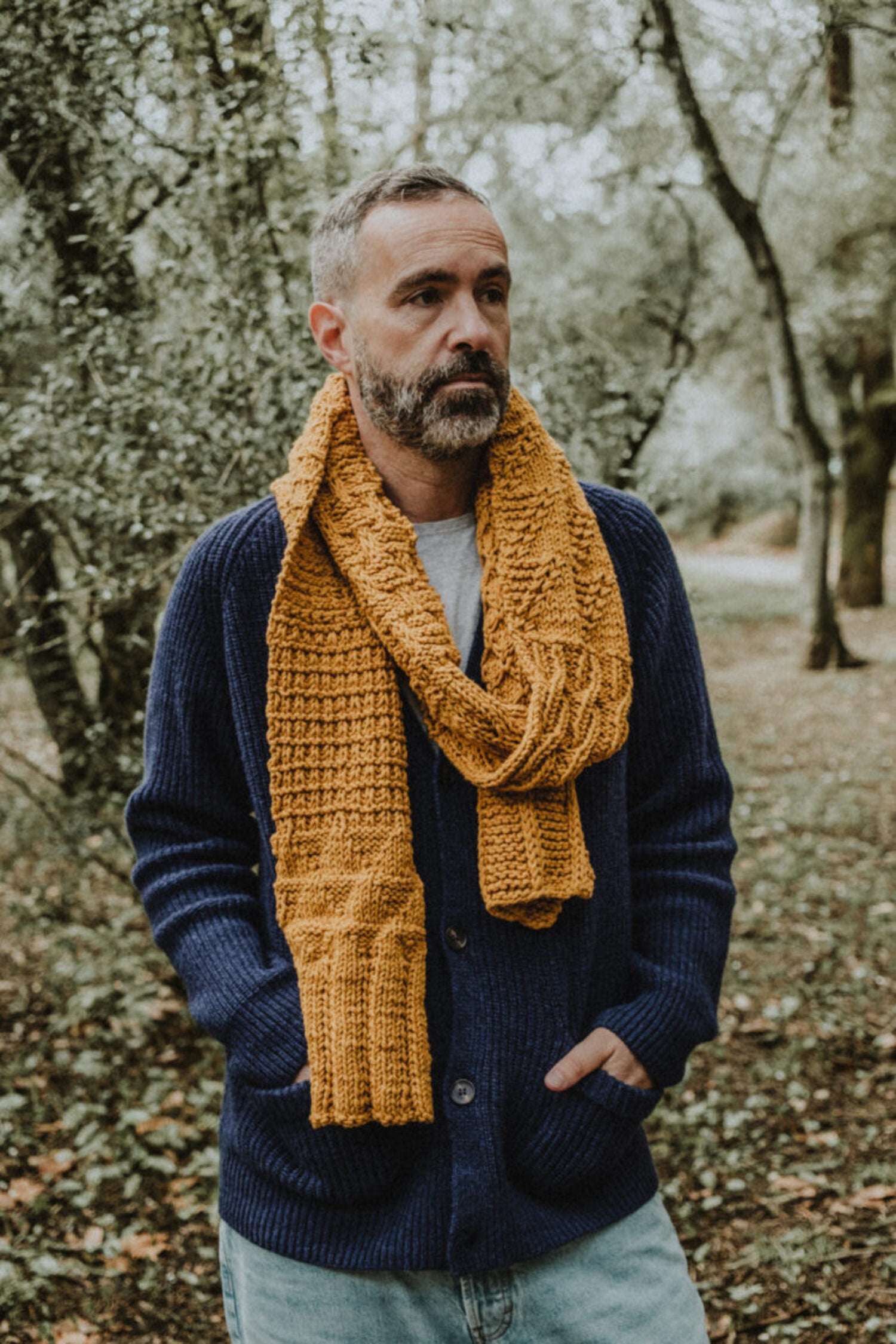 Bearded man wearing a mustard yellow handmade knit Re-Born Scarf paired with a dark blue cardigan in a natural forest setting.