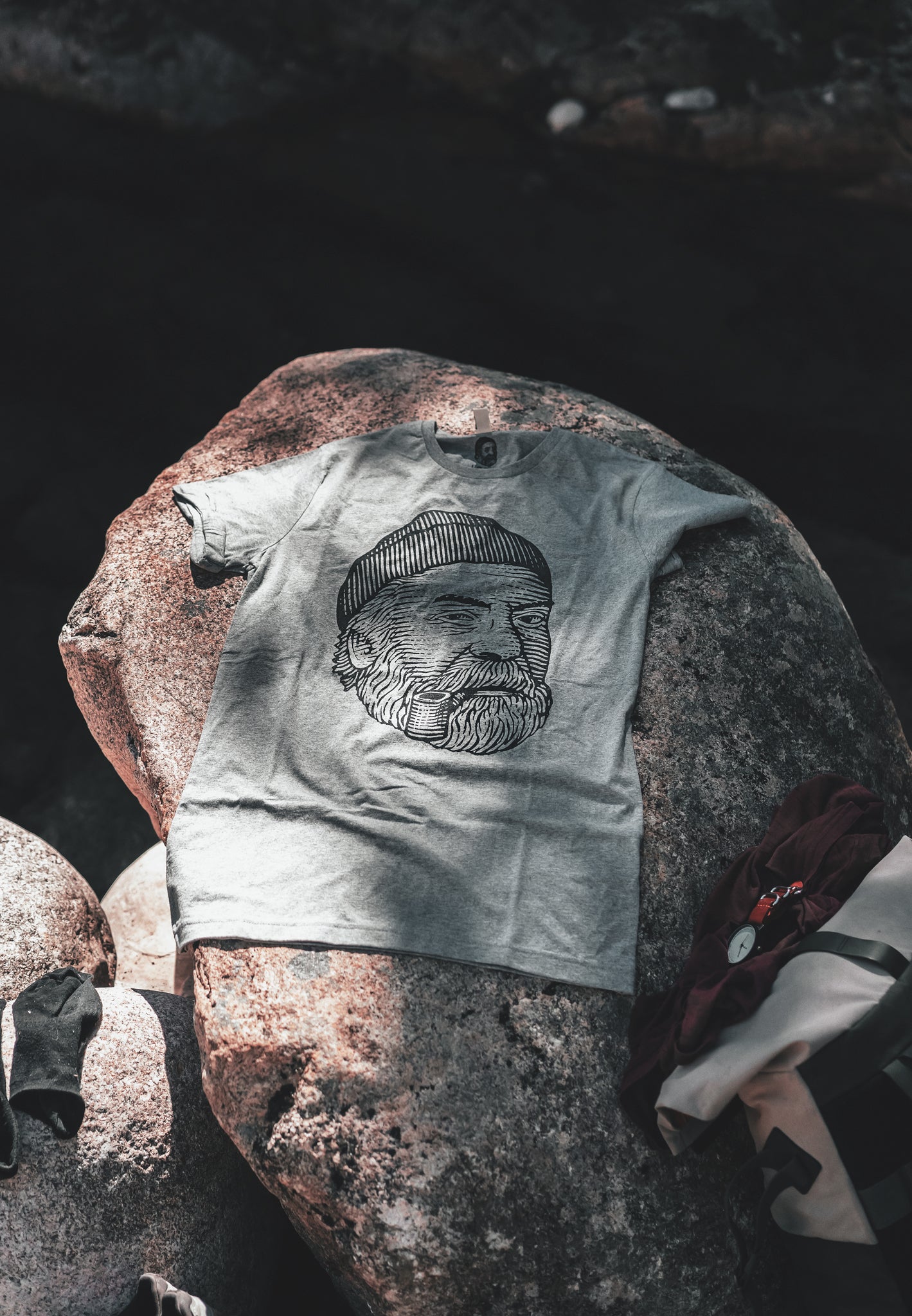 Grey LOBO t-shirt featuring the iconic logo of a bearded man with a beanie and pipe, placed on a rough granite rock in a natural outdoor setting, with sun and shadow.