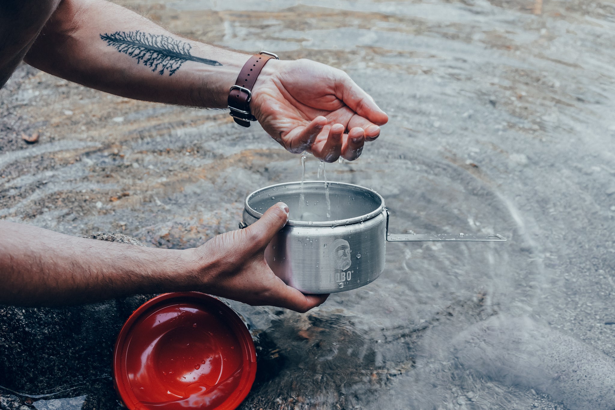 Close-up of hands washing a LOBO brand aluminum camping pot in a river. The pot features the logo of a bearded man with a beanie and pipe. The man has a pine tree tattoo on his forearm and wears a watch with a brown leather strap.