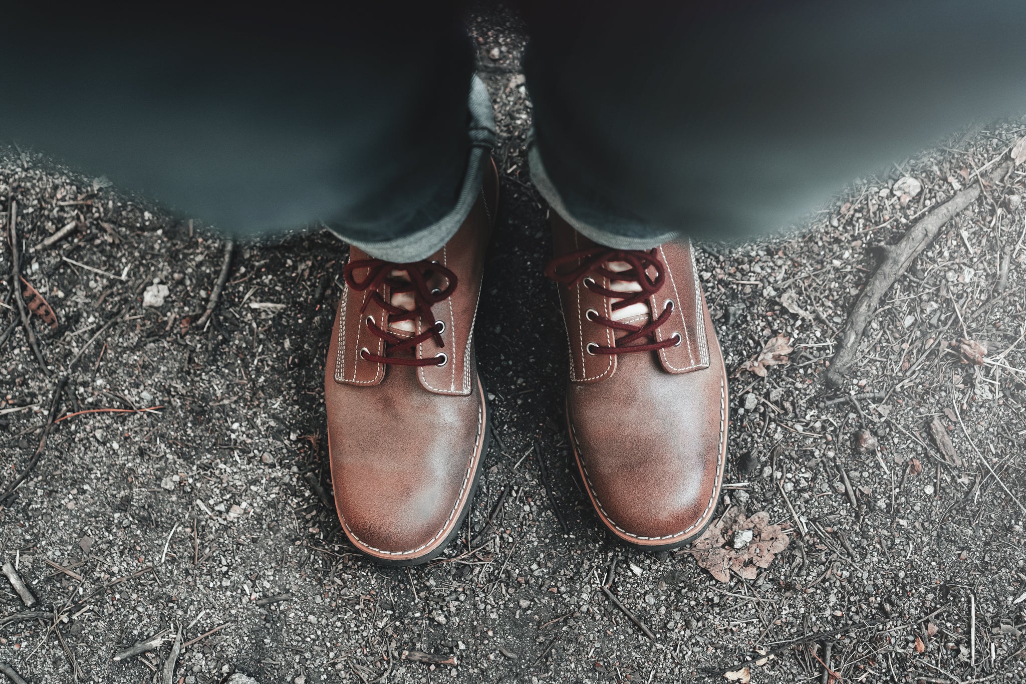 Top-down view (POV) of a wearer sporting the LOBO RAW Boots in brown leather with burgundy laces, standing on natural dirt and gravel ground, highlighting the rugged design and stitching details.