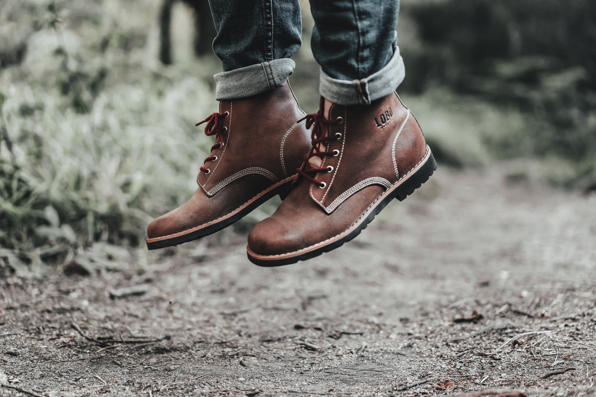 Legs with jeans and brown leather LOBO RAW Boots, captured in mid-air during a jump, with burgundy laces in motion, on a dirt trail in the forest.