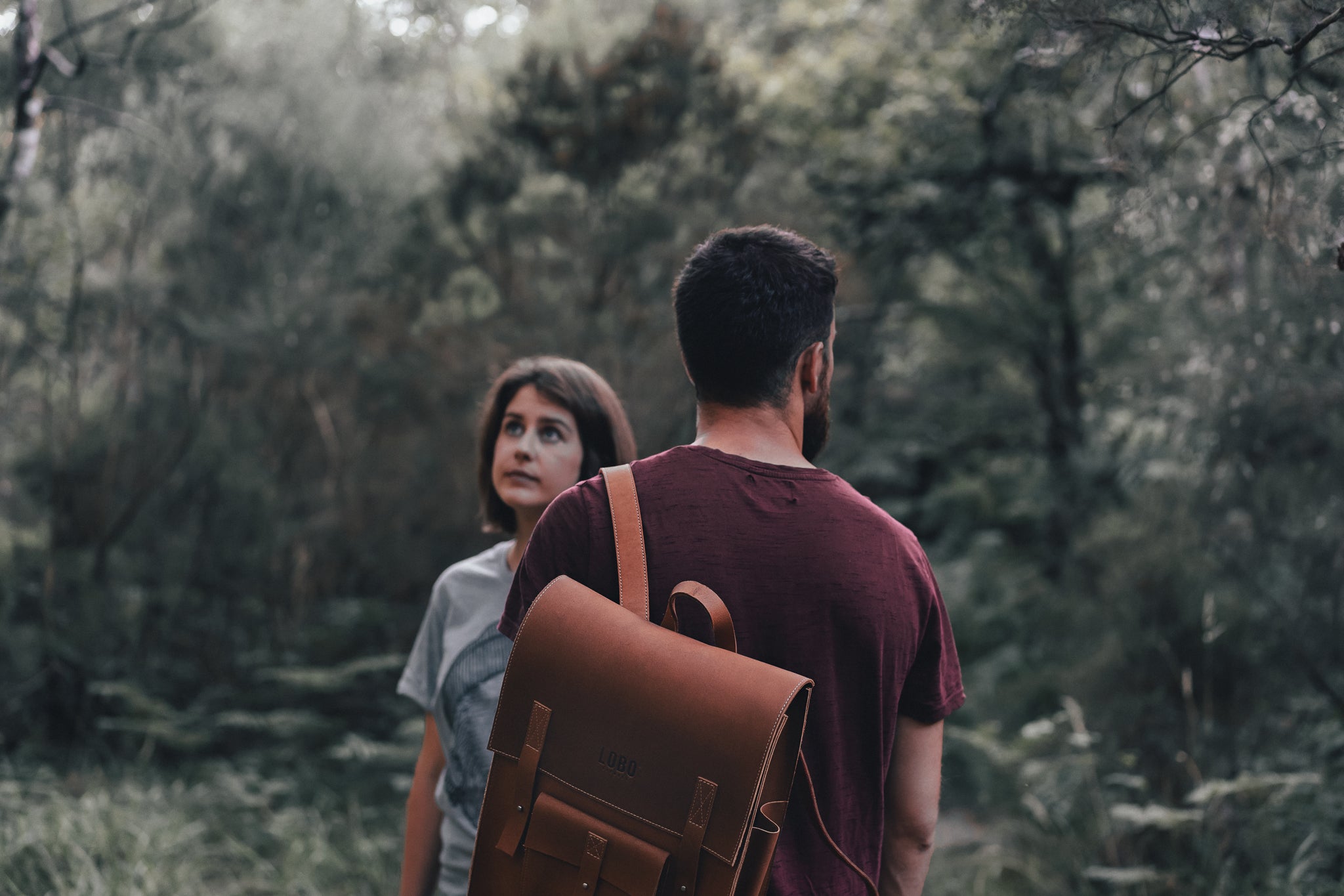 Man from behind wearing a LOBO brand brown leather backpack featuring the logo of a bearded man with a beanie and pipe, accompanied by a woman in a dense, natural forest setting.