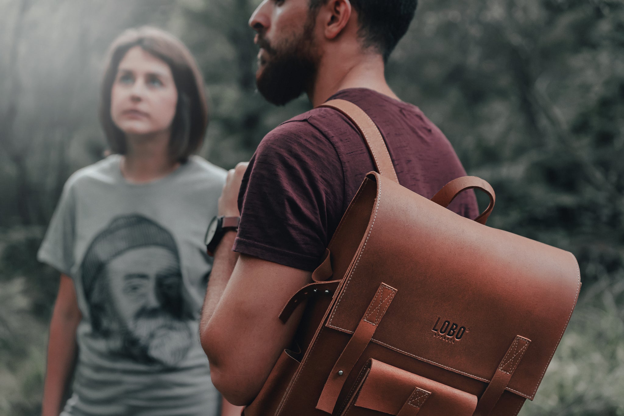 Close-up of a LOBO brand brown leather backpack with an engraved logo, worn by a man. In the background, a woman wears a grey t-shirt featuring the LOBO logo illustration: a bearded man with a beanie and pipe.