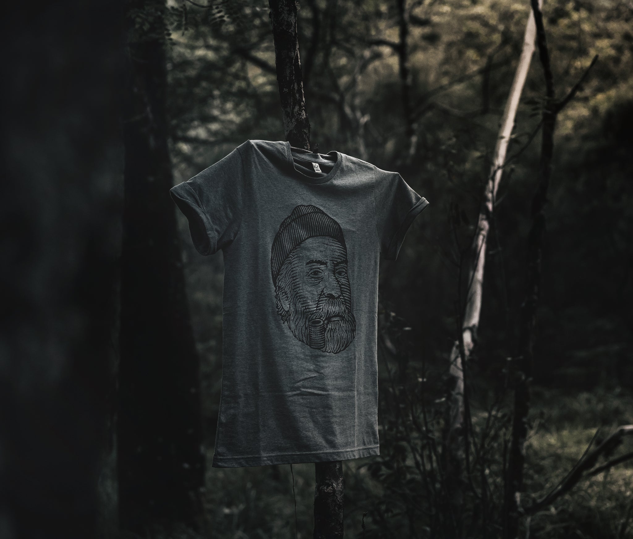 Grey LOBO t-shirt hanging on a tree trunk, showcasing the bearded man with a beanie and pipe print, in harmony with the organic forest setting.