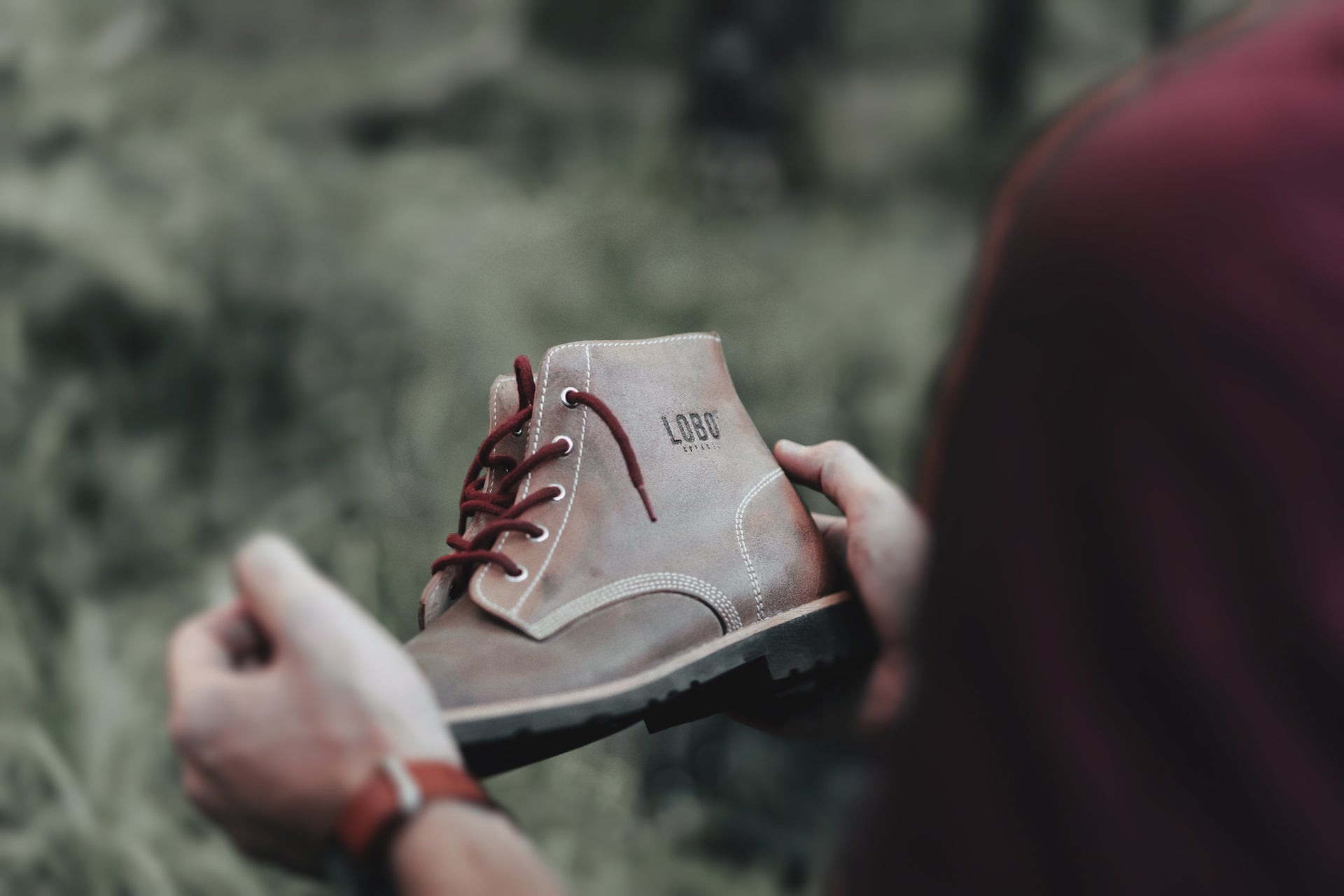 Close-up of hands holding the LOBO RAW Boots in brown leather, highlighting the craftsmanship and burgundy laces in a forest setting.
