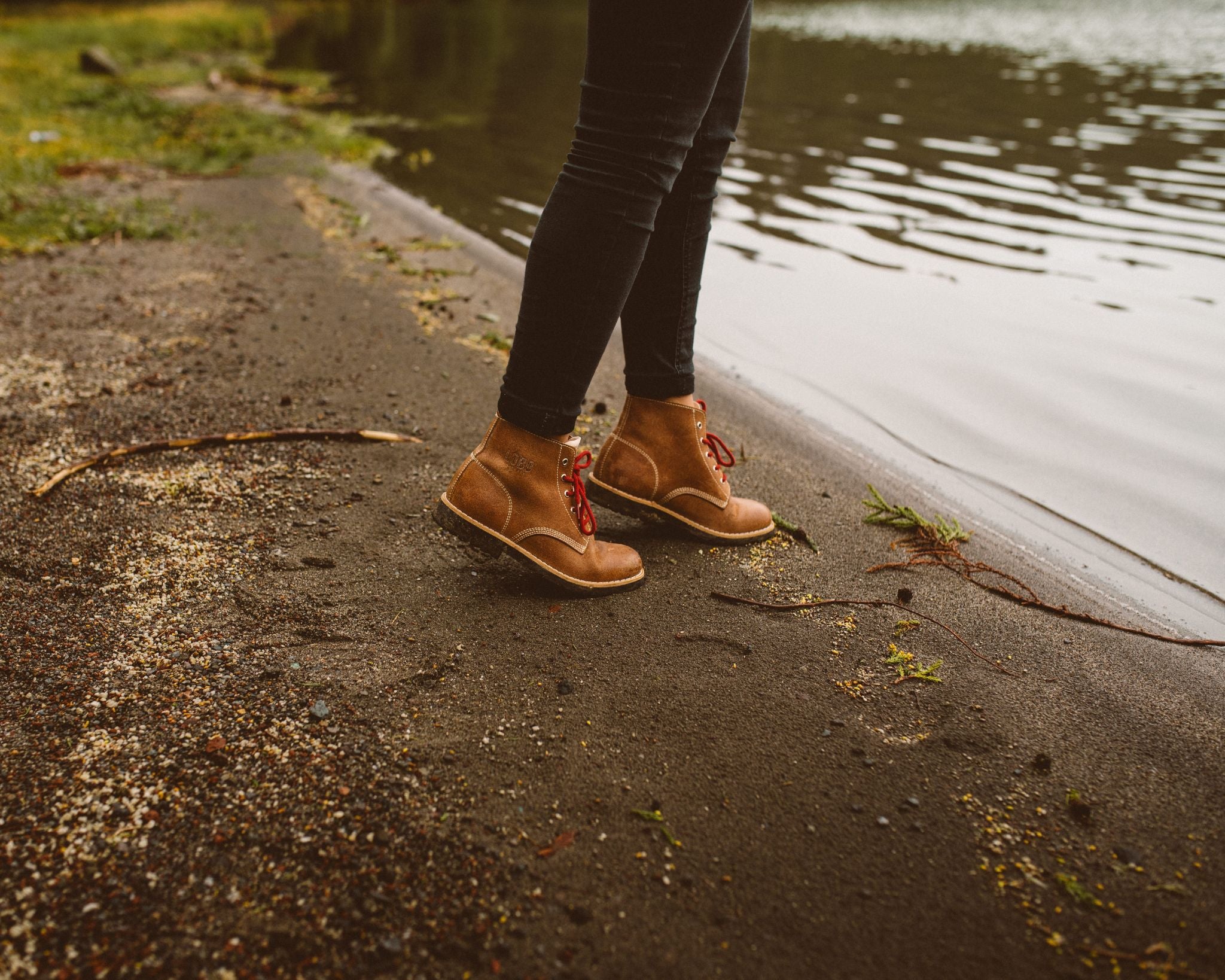 Close-up of brown leather Lobo Raw Boots on the shore of a lagoon with dark volcanic sand. The boots feature the engraved LOBO logo and red laces, showing their suitability for natural and wet terrains.