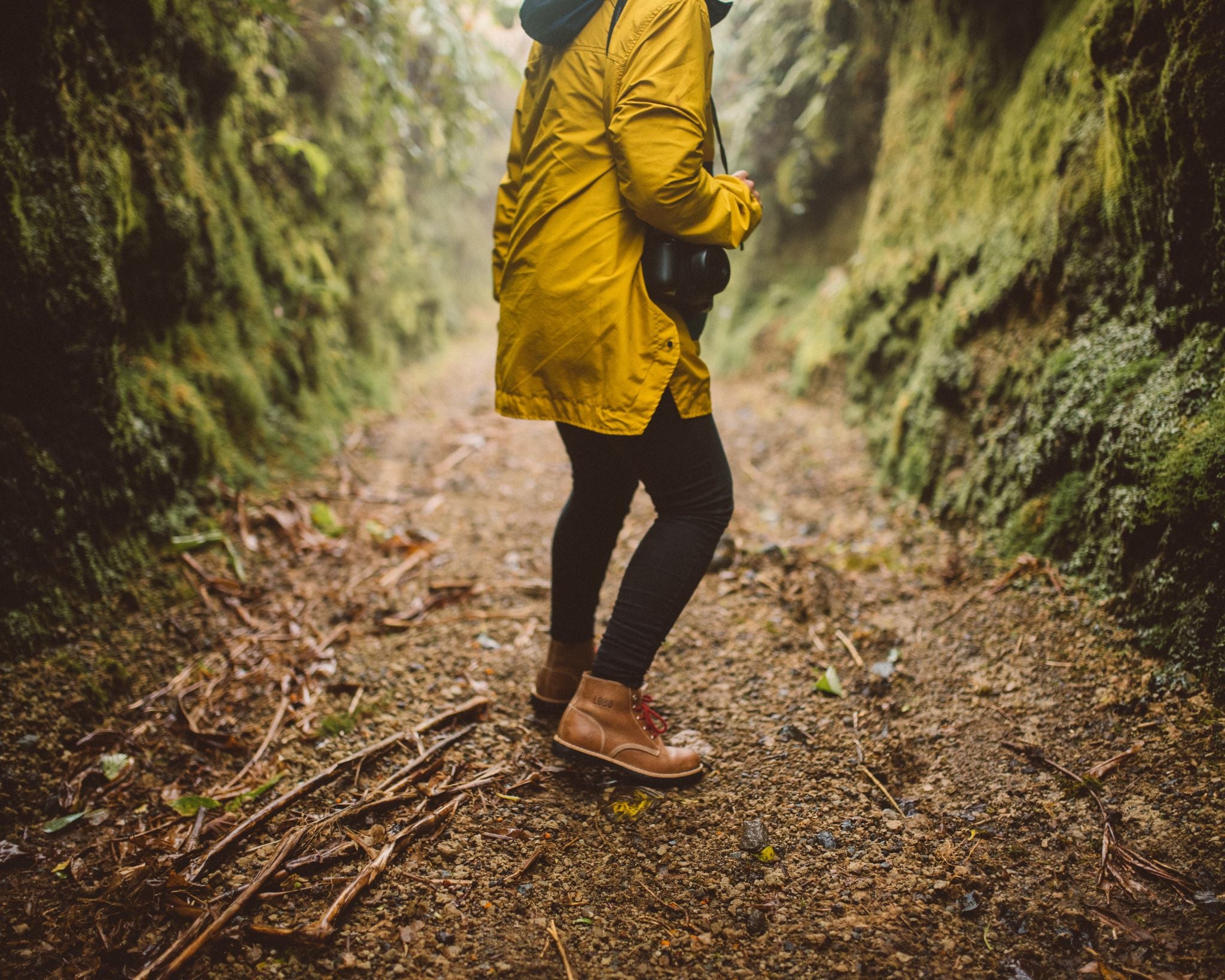 Person hiking through a damp, mossy forest trail wearing a yellow raincoat and brown leather Lobo Raw Boots. The boots stand out with their rugged design and red laces in an adventure setting.