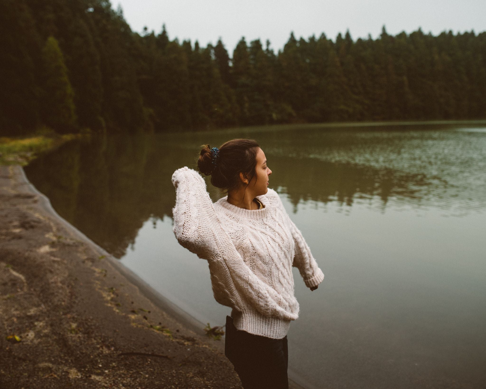 Woman wearing a chunky beige knit sweater and Lobo Raw Boots in a relaxed moment by the shore of Lagoa de São Brás, Azores. Serene nature setting with forest reflections on the water.
